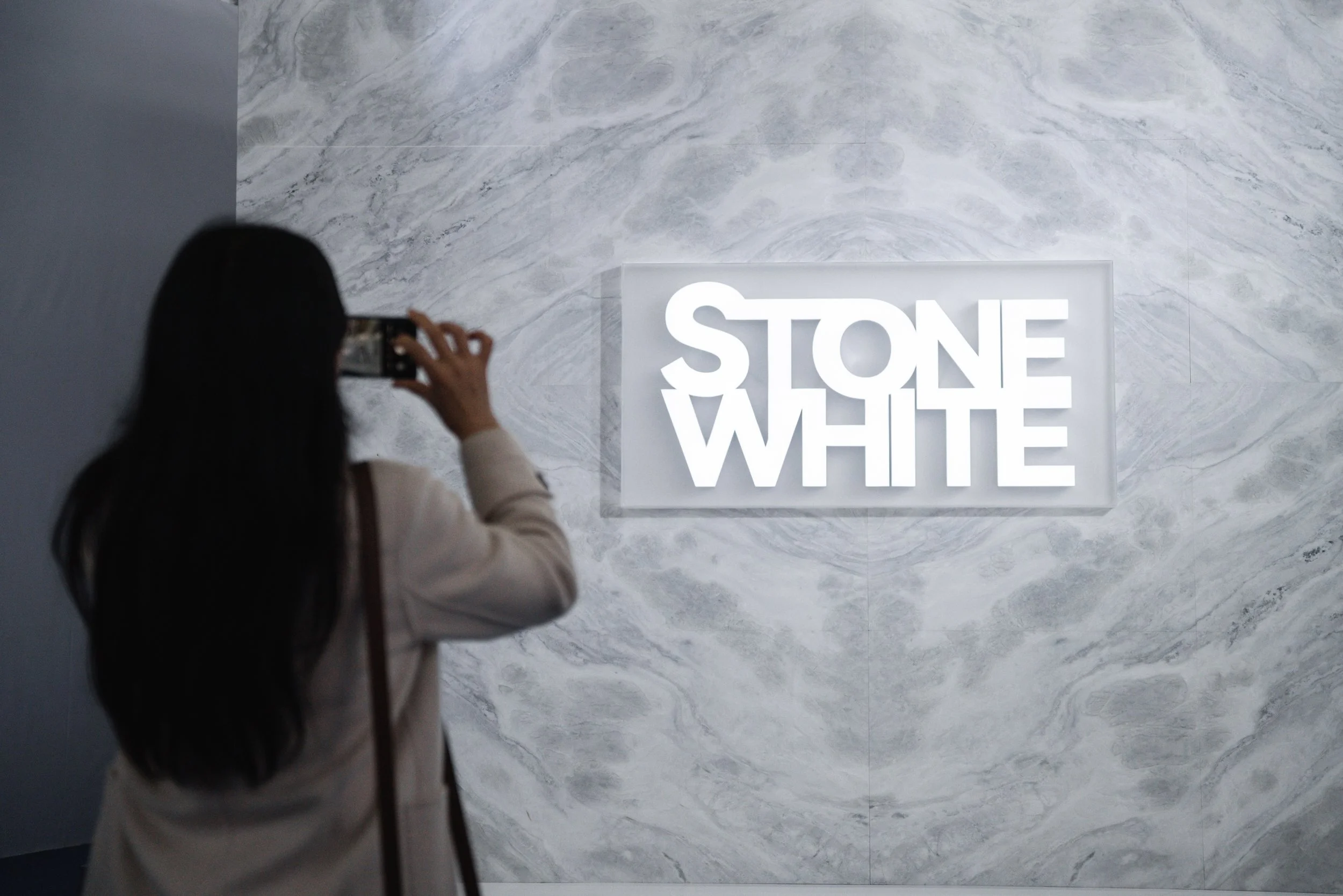 A woman with long dark hair taking a photo of a glowing sign that reads 'STONE WHITE' on a gray, marbled wall.
