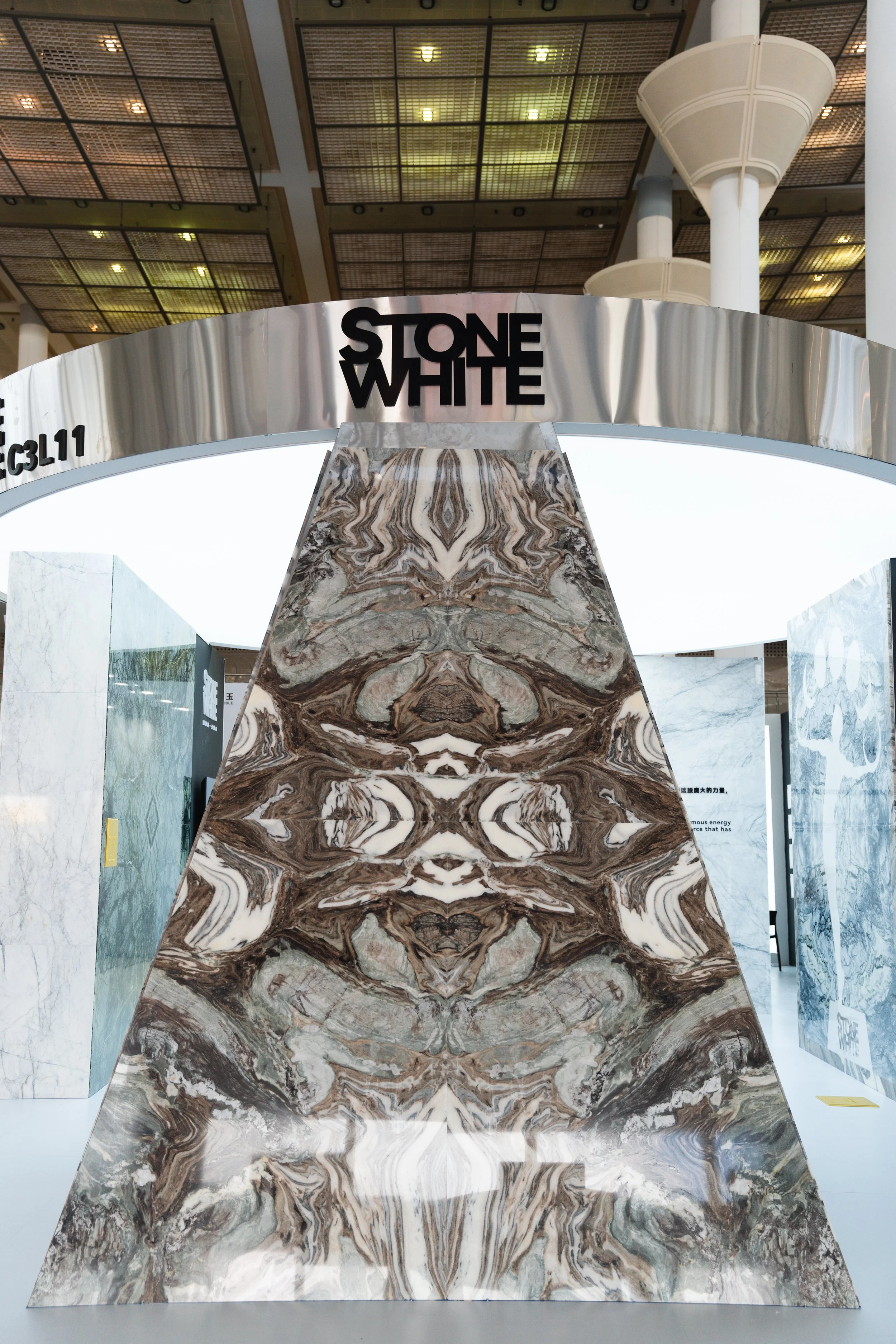 Exhibit display at Stone White featuring a large, mirrored marble slab with symmetrical, intricate brown, white, and gray veining. The exhibit is located under a modern, high ceiling with metal grates and white columns.