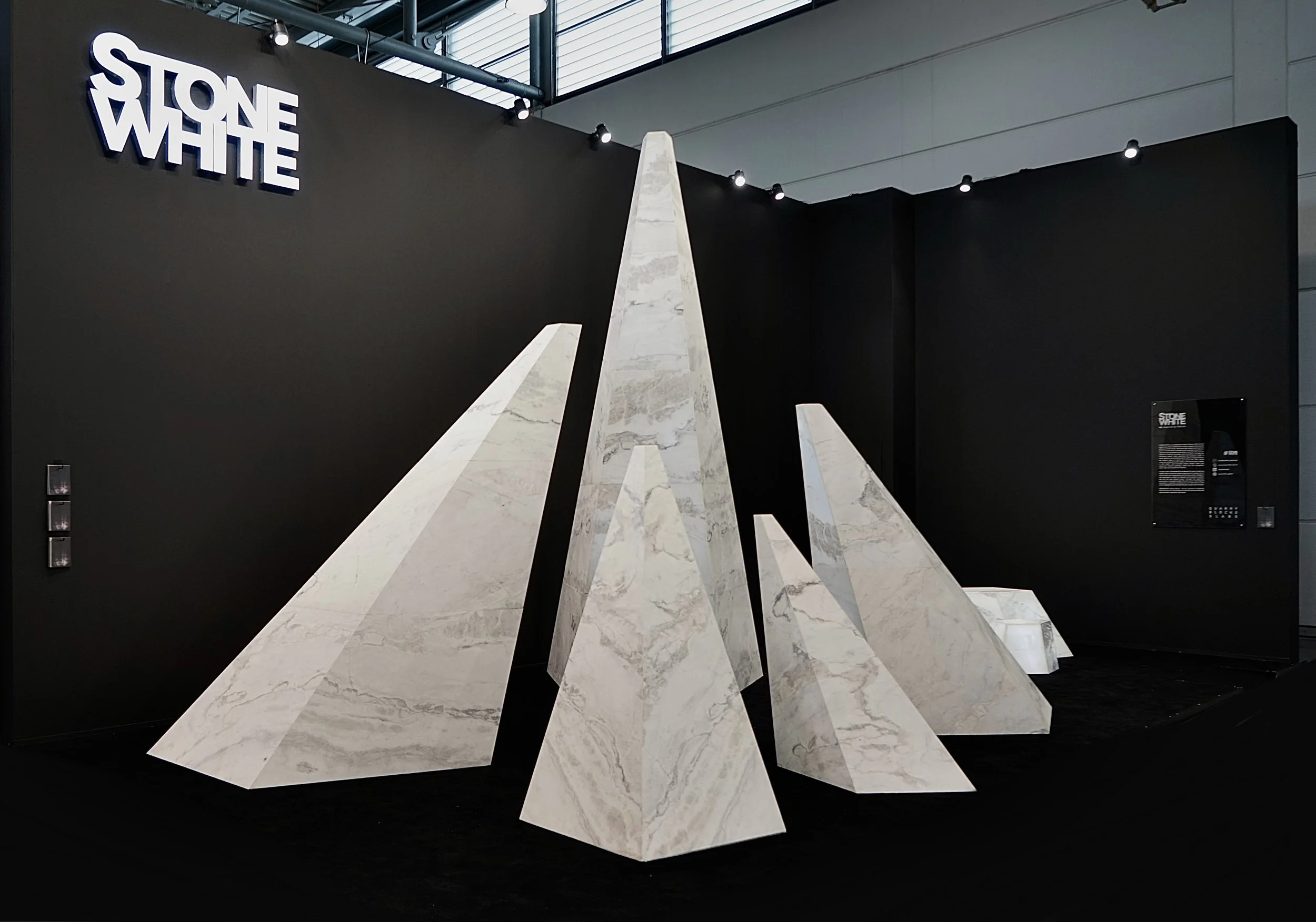 Art installation featuring tall, angular, white marble-like geometric sculptures resembling mountains, set against a black backdrop with the words 'STONE WHITE' displayed on the wall.