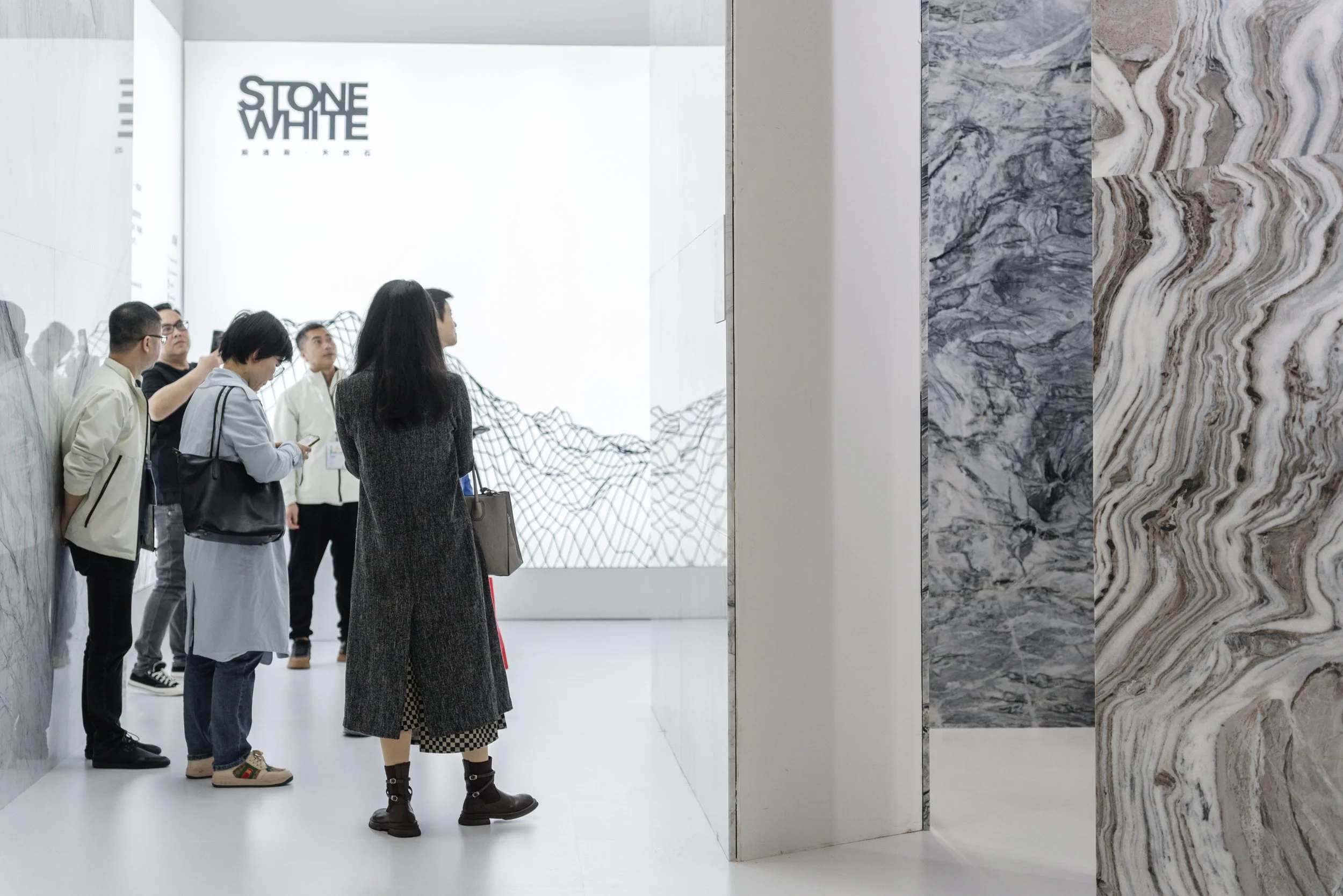 A group of six people are standing in an art gallery, observing a white art piece that has the words 'Stone White Market' written on it. The gallery has a white floor and walls, with large marble-like panels on the right side of the image.