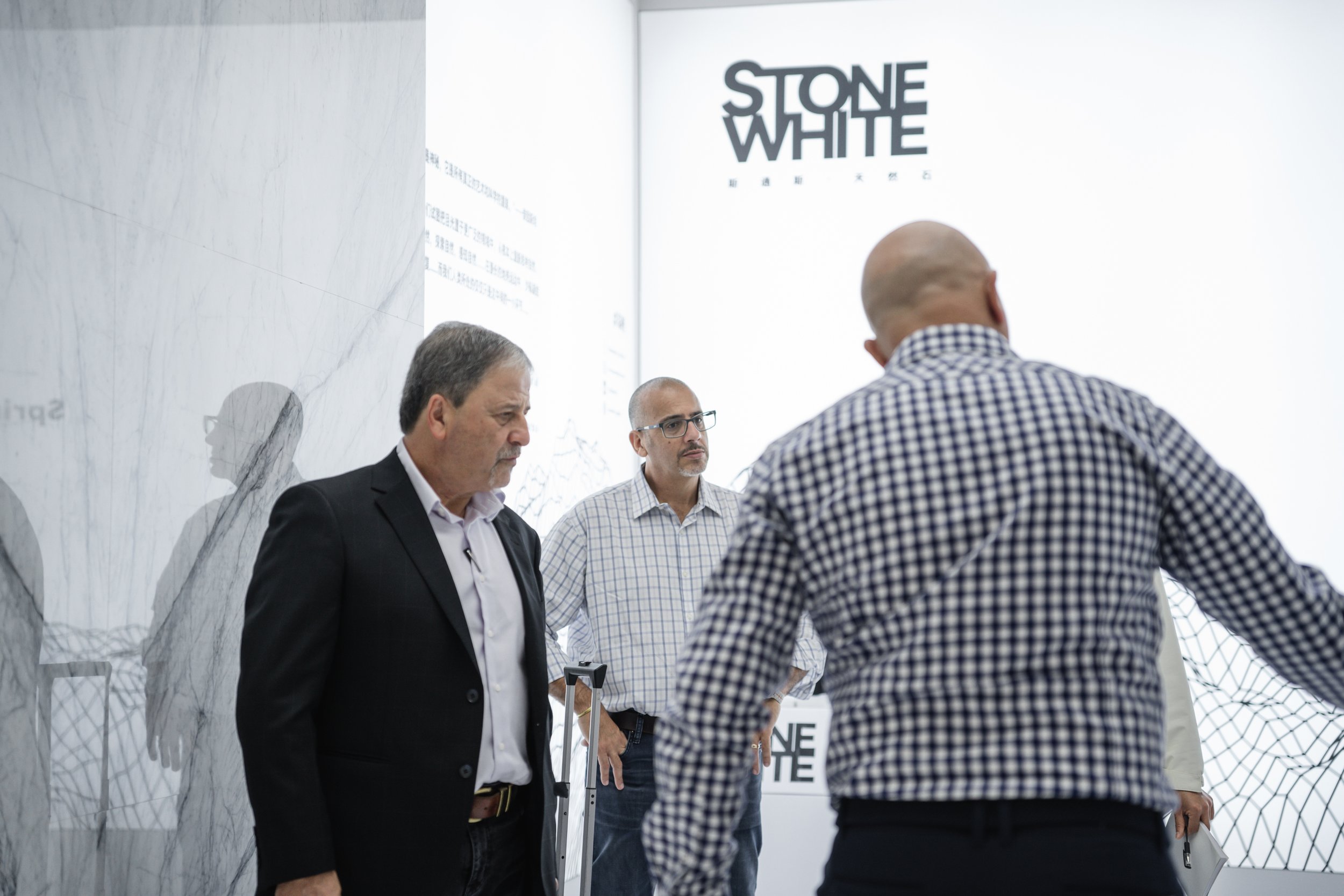 Three men standing in a room with a modern design. Two of them are facing a third man who has his back to the camera. The background features a large white wall with text that reads 'STONE WHITE' and some smaller text underneath.