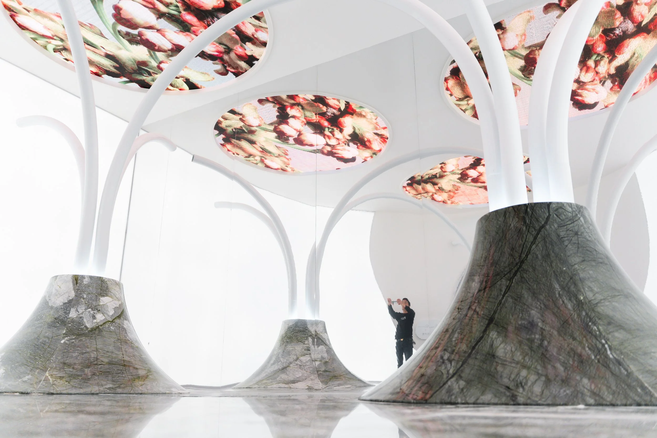 Modern art installation featuring large tree-like white structures with wide bases resembling rocks, with circular ceiling panels displaying floral patterns and a person taking a photo in front of a reflective surface.