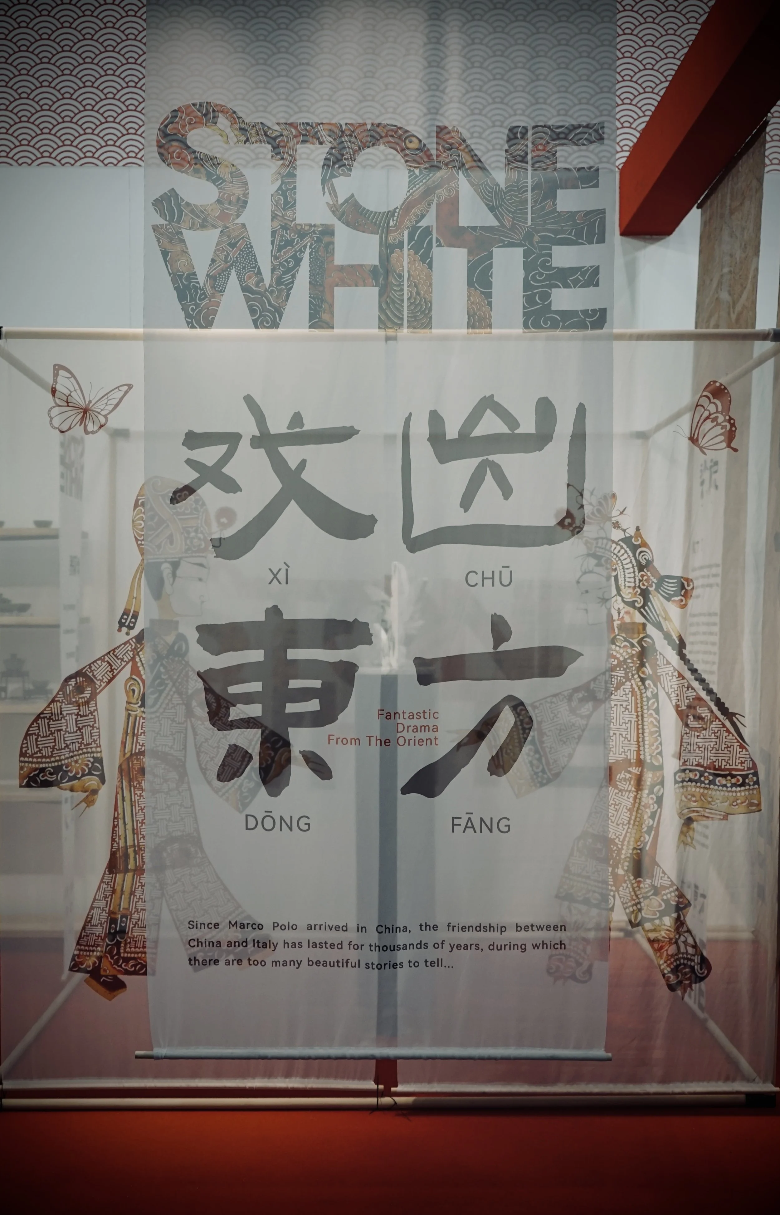 A display board with Chinese characters and the phrase 'Fantastic Drama from the Orient' and a paragraph about Marco Polo's arrival in China, featuring illustrations of traditional Chinese figures and butterflies.