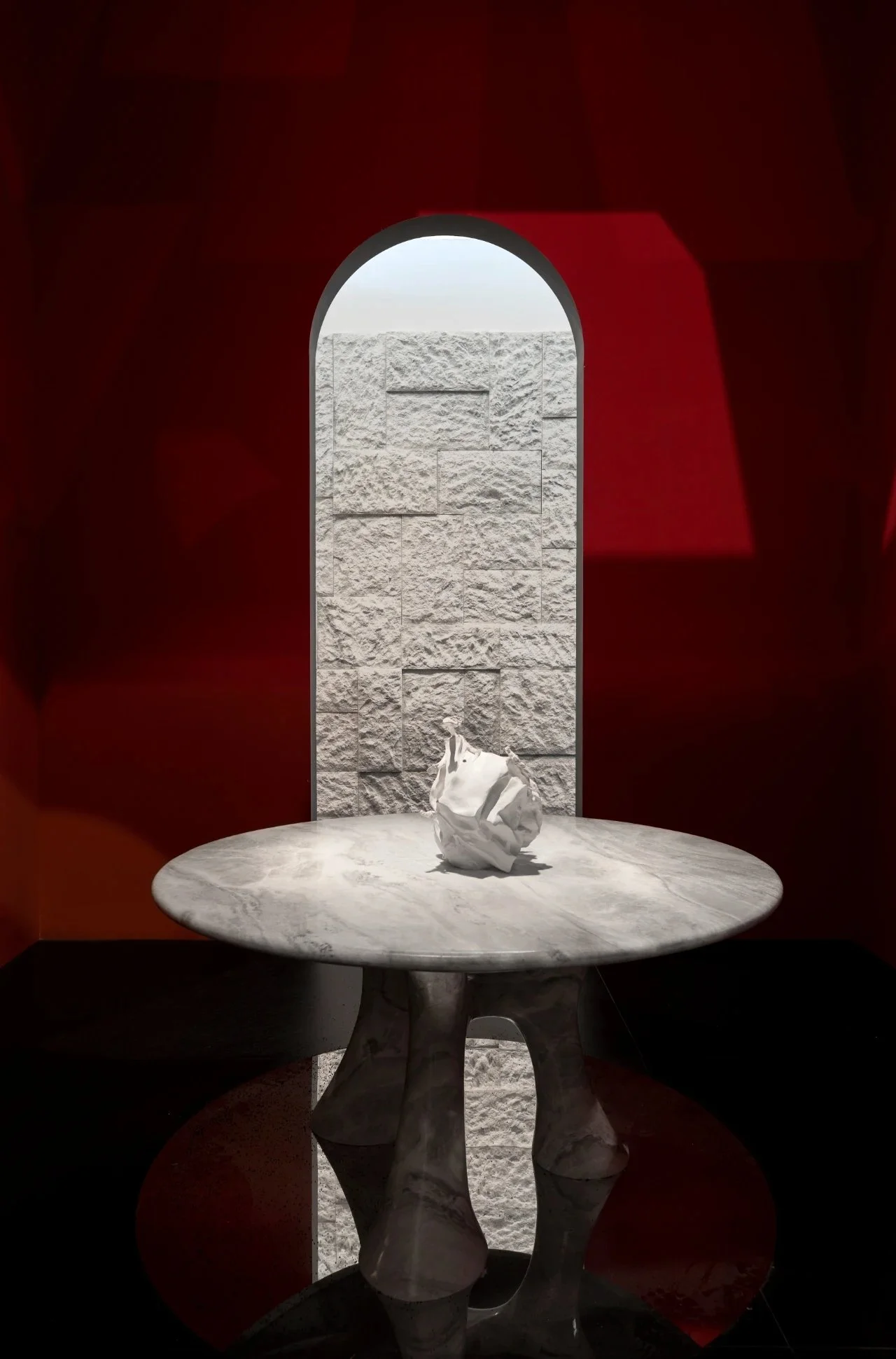 A round marble table with a crumpled white object on top, placed in front of a red wall with a narrow arched window revealing textured stone bricks outside.