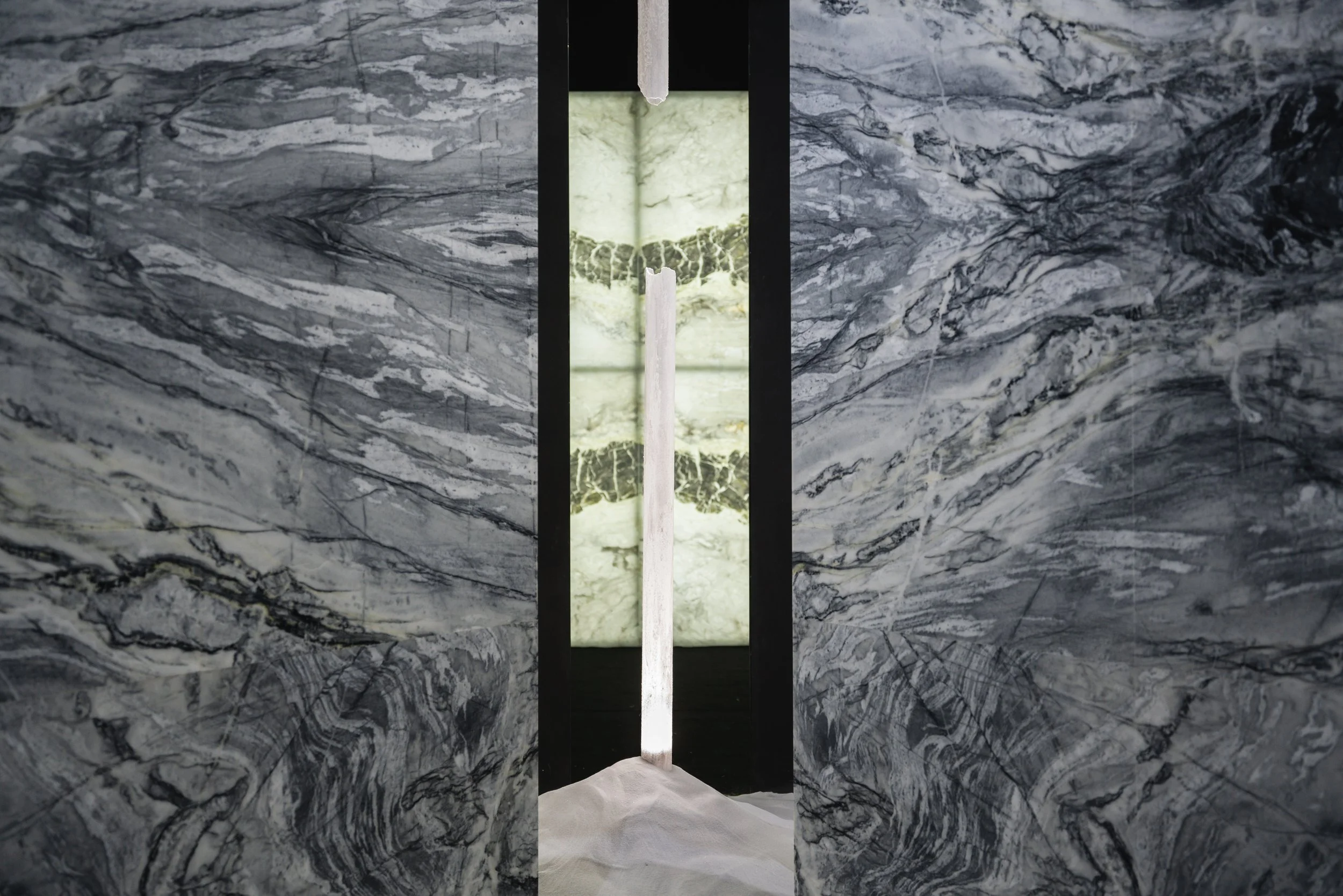 A narrow slit between two dark, marbled stone walls, revealing a glowing green stone panel with black veining in the background, and a white vertical object in the foreground.