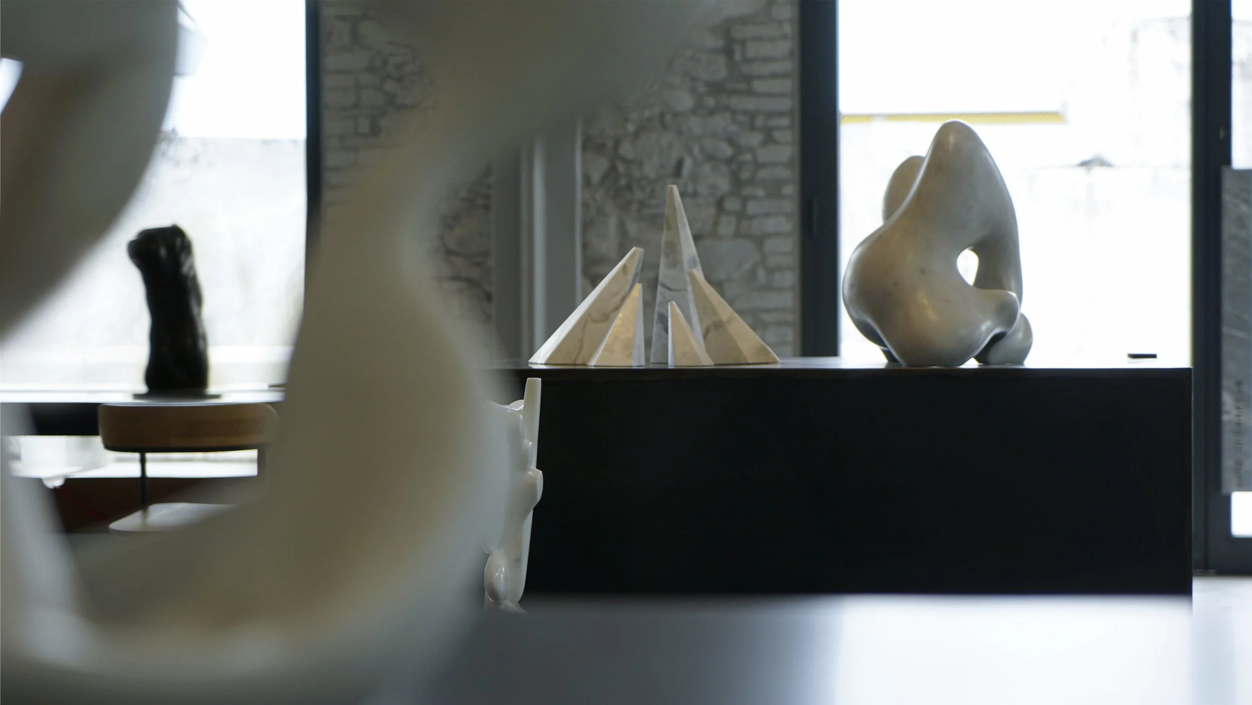 Interior of an art gallery displaying abstract sculptures and geometric artworks on a black surface, with brick wall and large windows in the background.