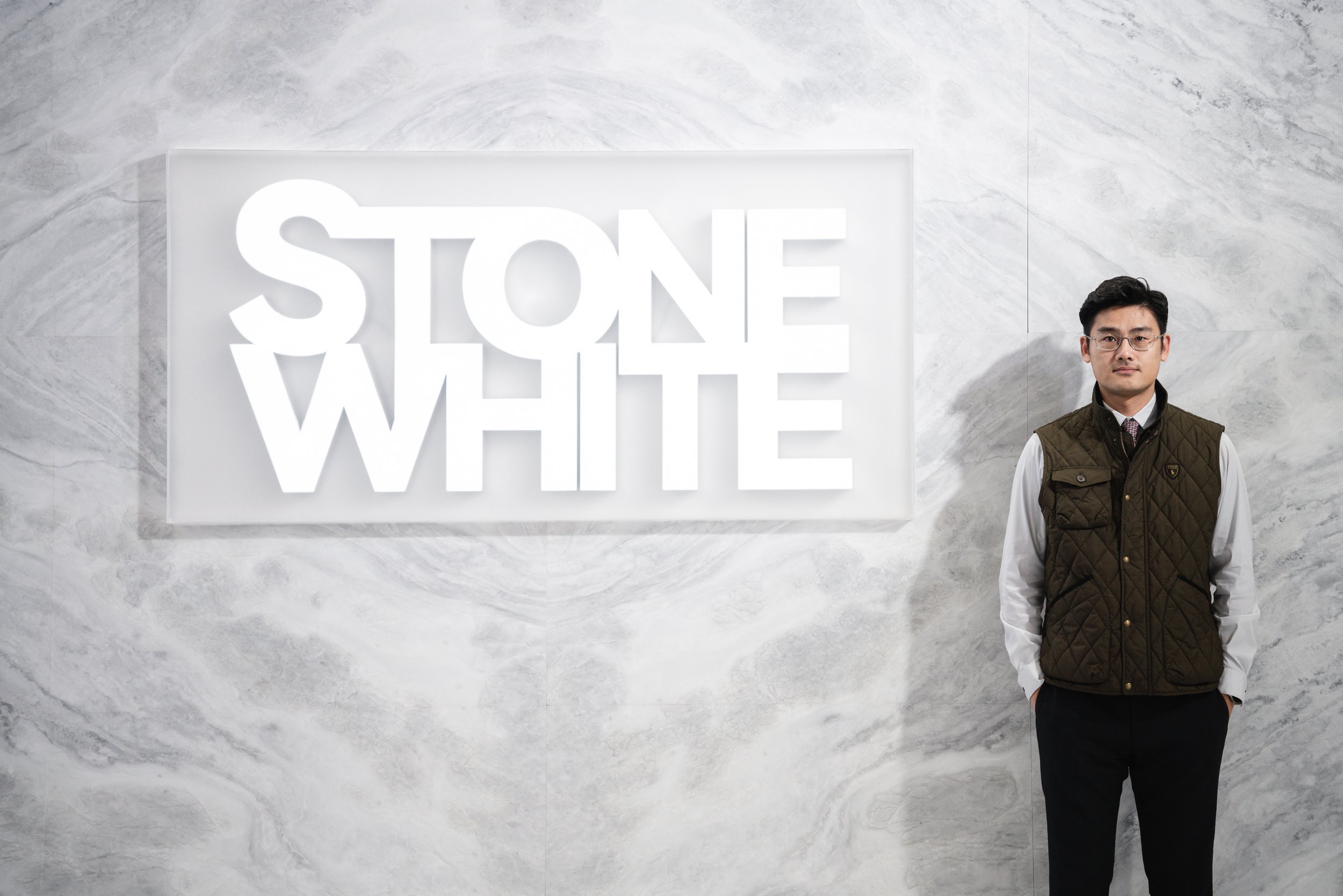 A man in glasses and formal attire, including a white shirt and dark pants, stands with hands in pockets in front of a marble wall. To his left, a large illuminated sign displays the words 'STONE WHITE' in bold, white letters.