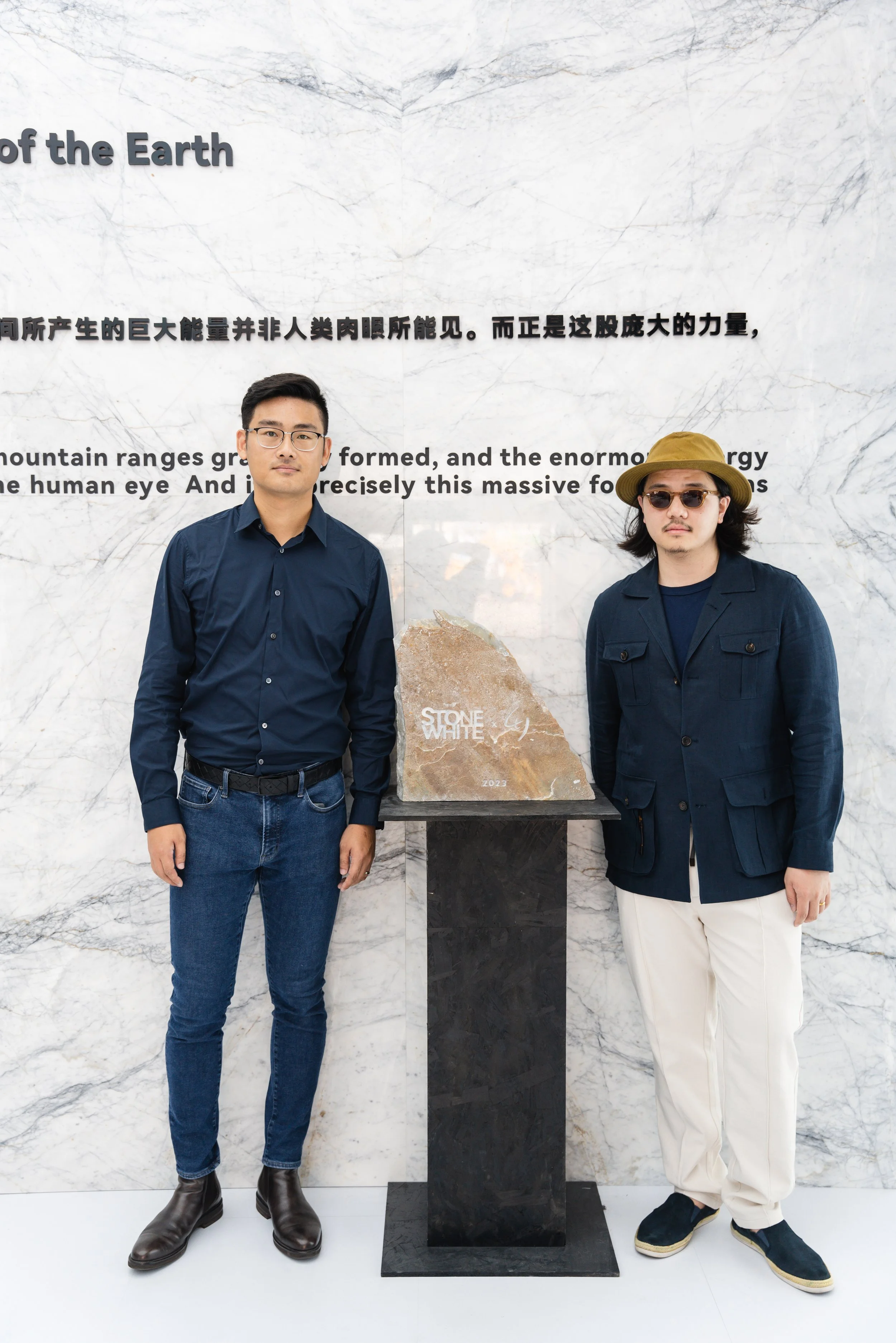 Two men standing next to a stone on a pedestal inside a building with a marble wall background that has text in Chinese and English.