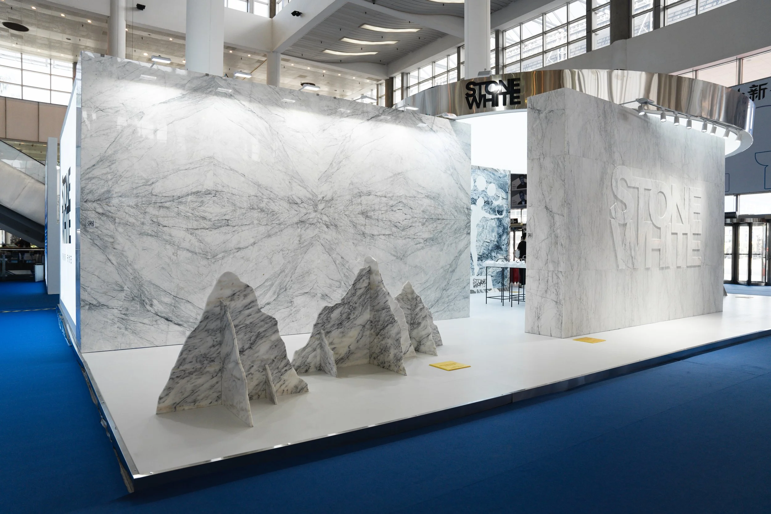 Exhibition booth with marble-themed displays, including a large marble wall, smaller marble sculptures resembling mountains, and a panel with the words 'STONE WHITE' engraved, set in an indoor event space with high glass ceilings.