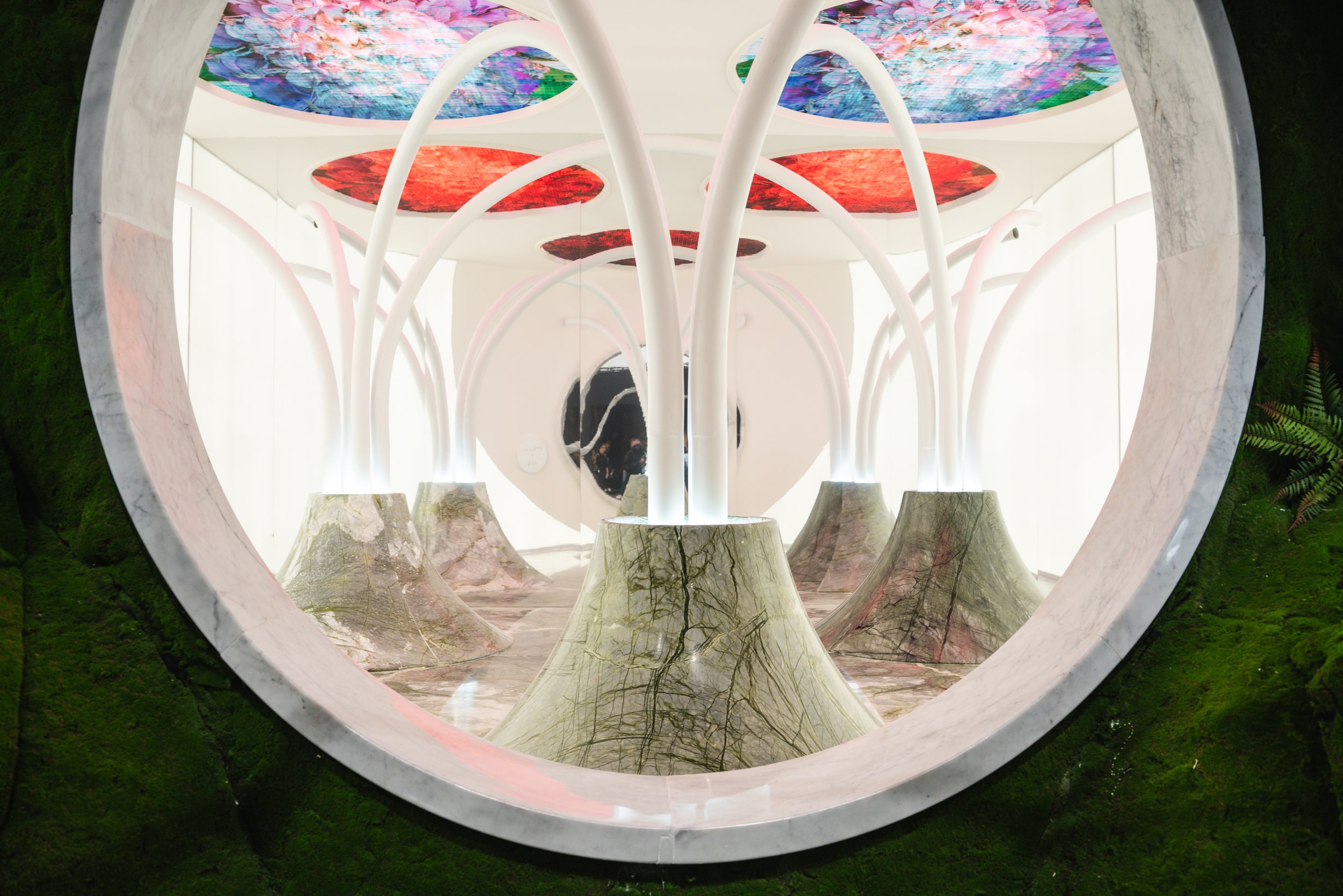 Interior view of an art installation featuring tree stump sculptures with smooth, polished surfaces and white, curved supporting branches extending to colorful, patterned circular panels on the ceiling. The scene is framed by a large, rounded opening