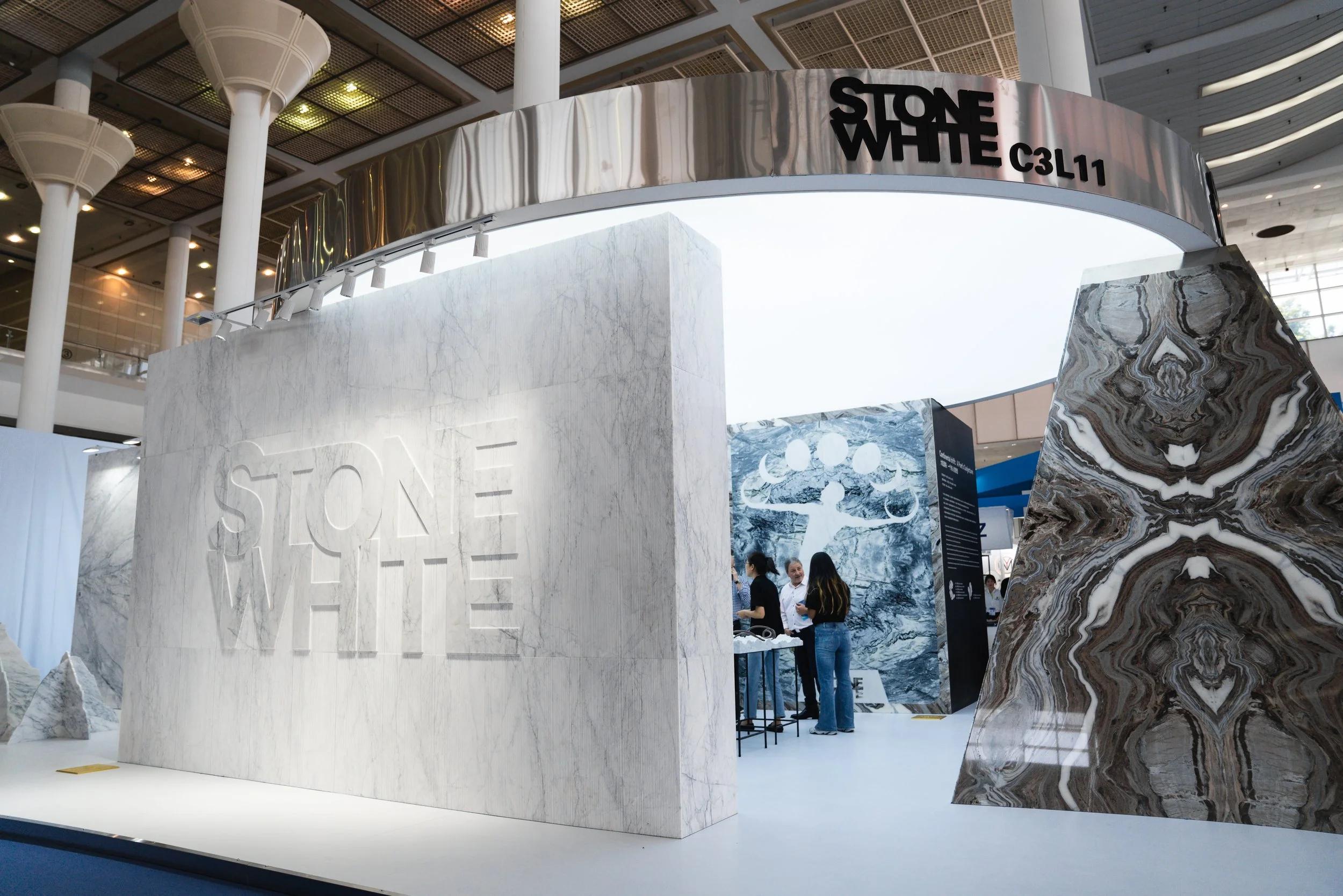 Trade show booth displaying various marble slabs labeled 'Stone White,' with a large branding sign 'Stone White' and people interacting inside the booth.