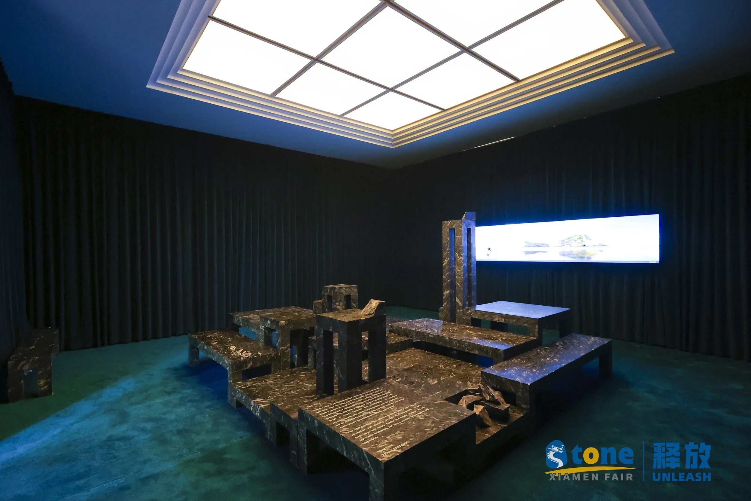 A modern room with black curtains, a large illuminated ceiling panel, and textured dark stone furniture including benches and a table, with a bright digital screen on the wall displaying a seaside landscape.