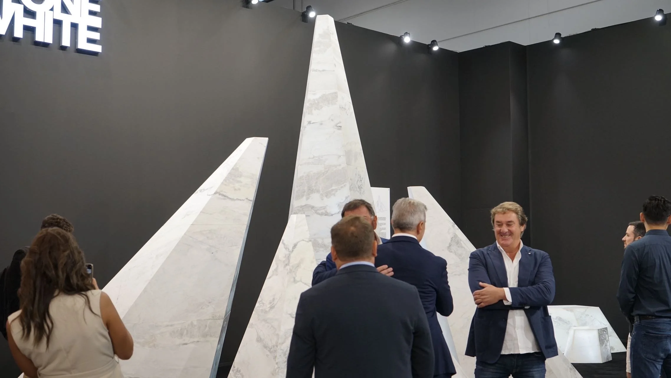 Group of people in business attire standing and talking in front of large white marble slabs at an exhibition or trade show.