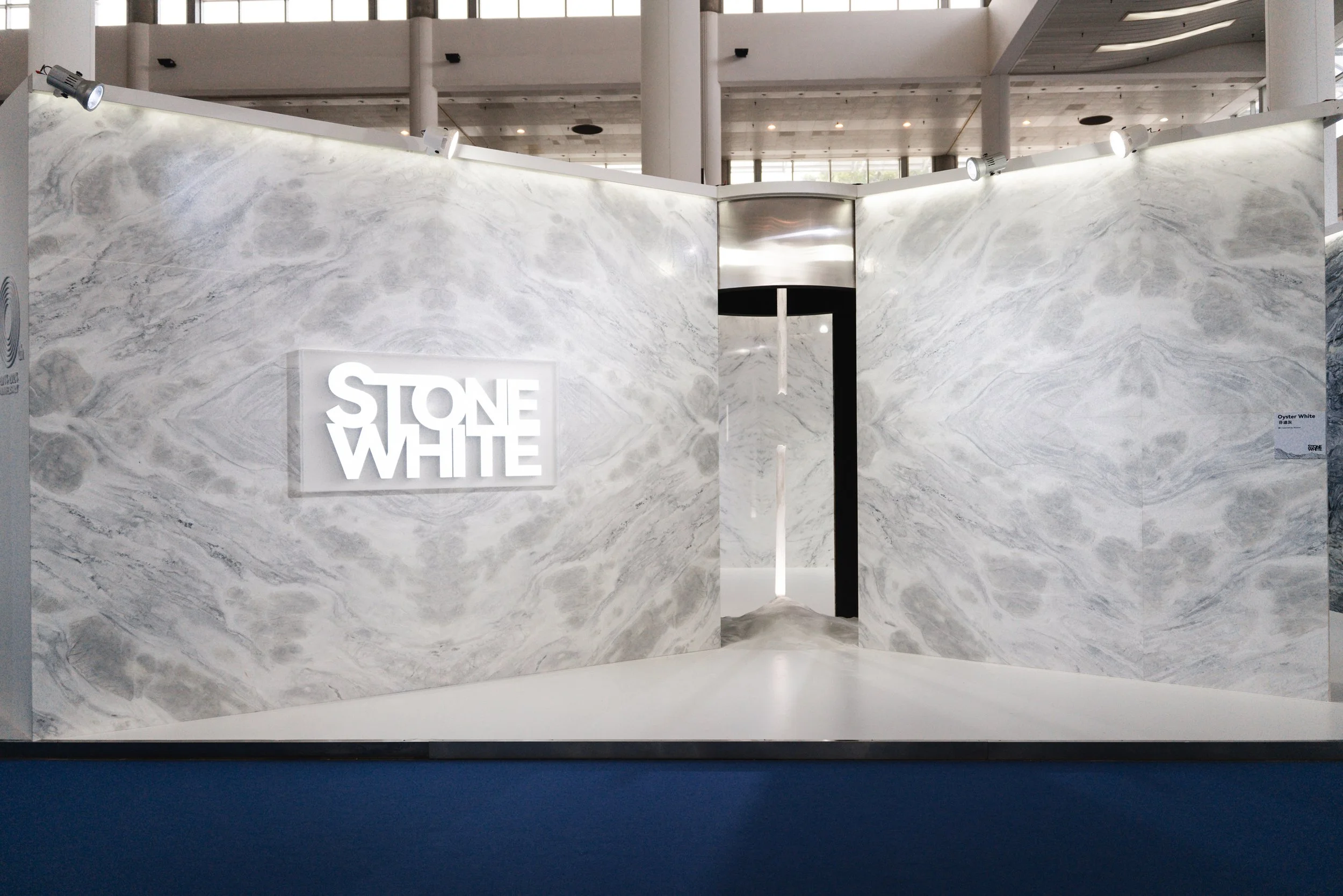Exhibition booth with marble walls and a illuminated sign reading 'STONE WHITE,' located in a large open indoor space.