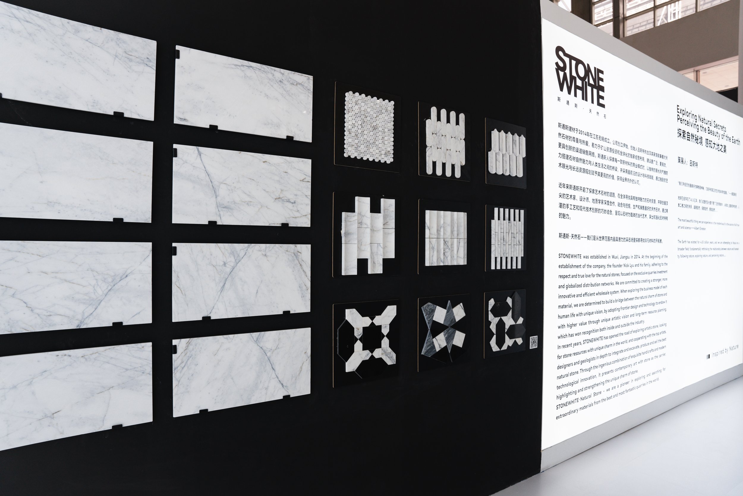 Display wall with panels of white marble and geometric pattern samples, with an adjacent informational panel about Stone White, a company specializing in natural stone materials.
