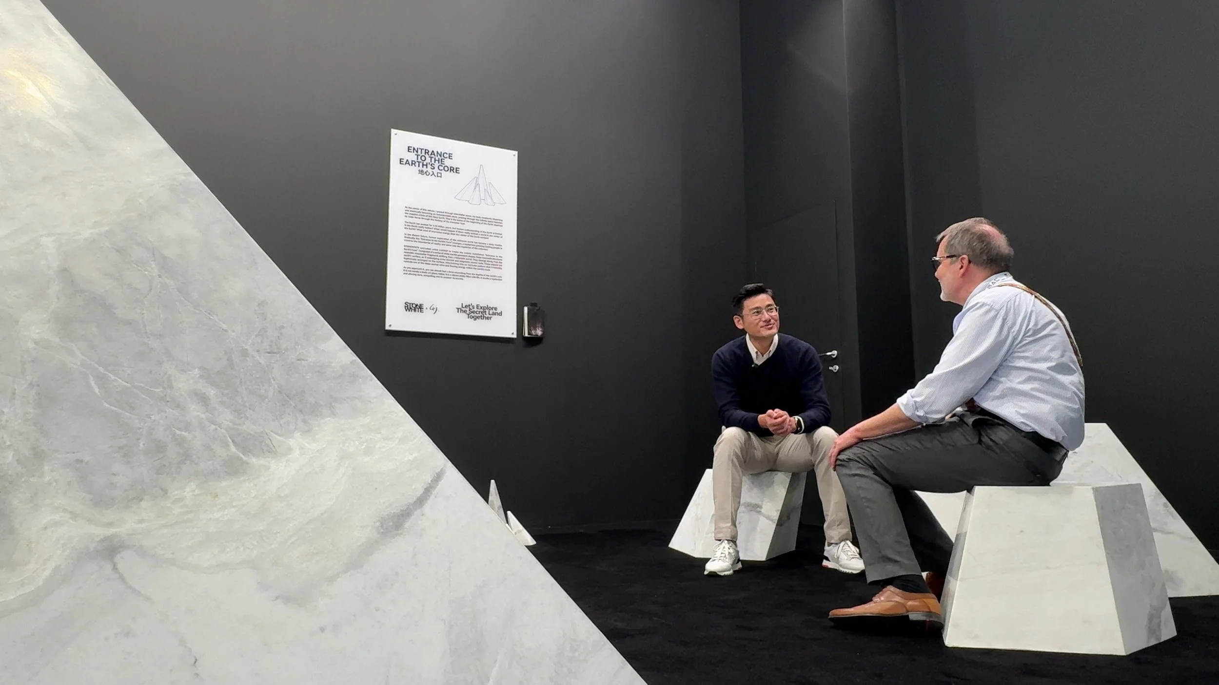 Two men sitting on geometric white benches in a black-walled room, engaged in conversation. One man is Asian, wearing a navy sweater and beige pocket, the other is Caucasian, wearing a white shirt and dark gray pants. A poster about Earth's core is o