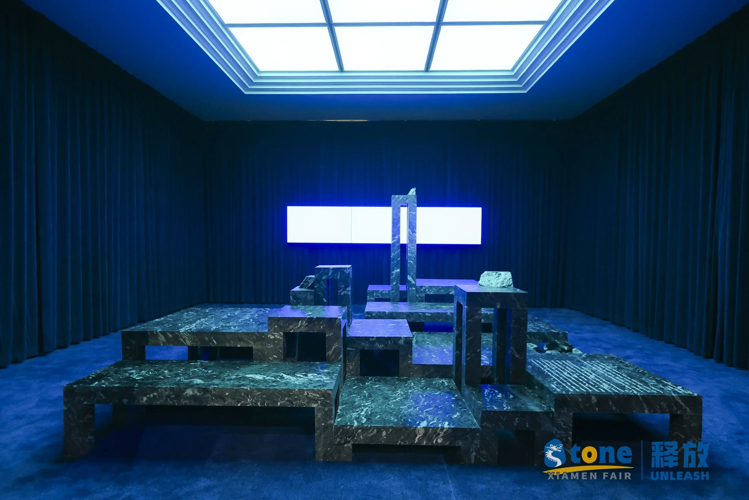Modern installation with blue lighting, marble geometric structures, illuminated rectangular screen, and curtains in the background at Xiamen Fair.