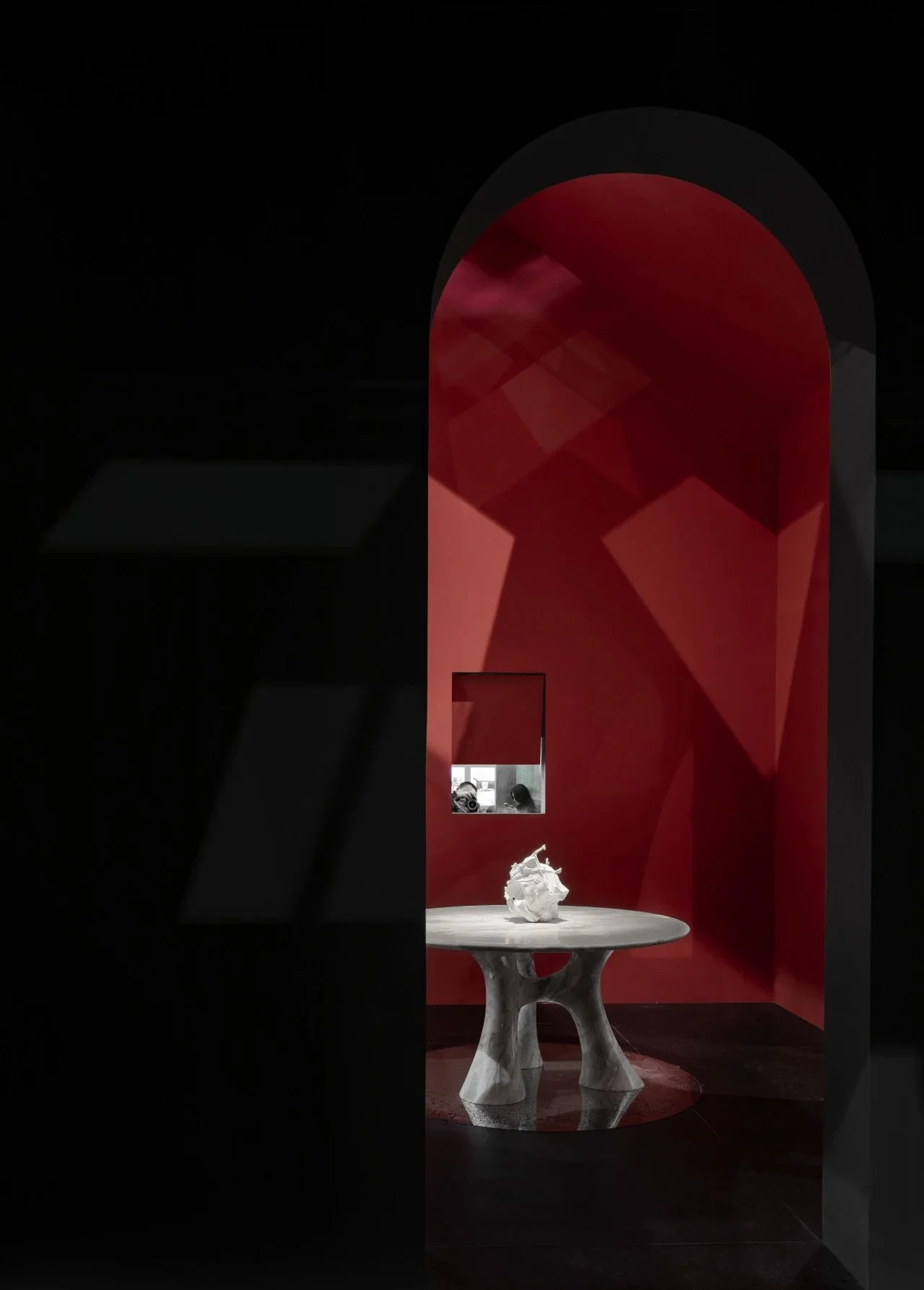 An interior view of a room with red walls and a semicircular archway, featuring a white sculpture on a round wooden table and a small window in the background showing two people.