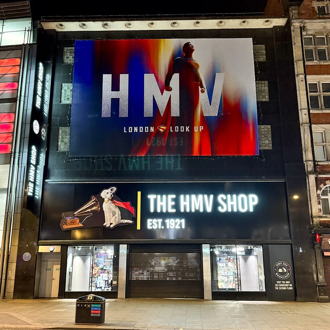 Billboard advertising HMV store with a Superman-themed poster saying "LONDON LOOK UP" and a storefront featuring a dog wearing a red scarf with a gramophone, and the sign "THE HMV SHOP EST. 1921".