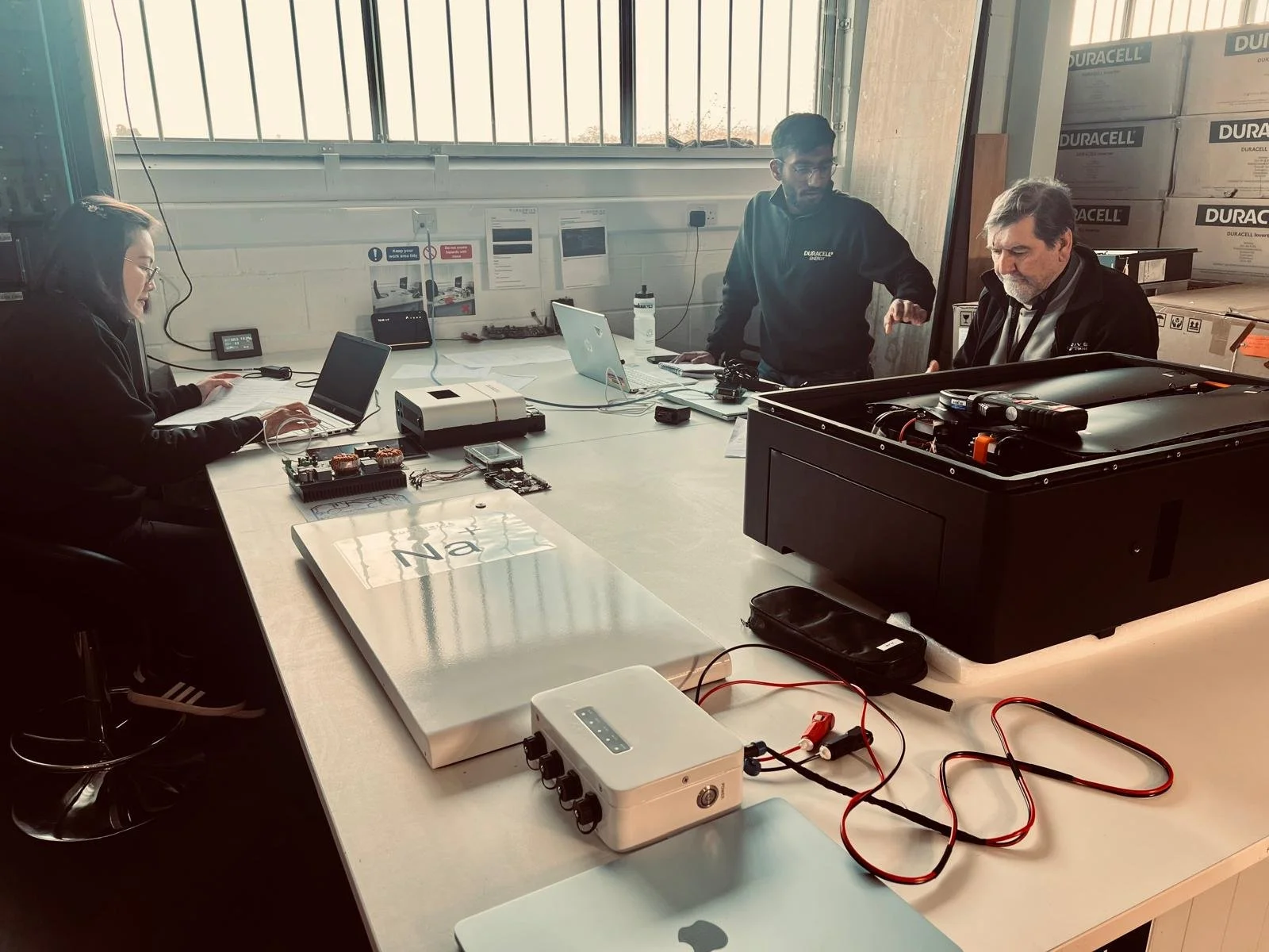 Three Product development people working at a large table in a workshop, with solar battery equipment and development products on the table.