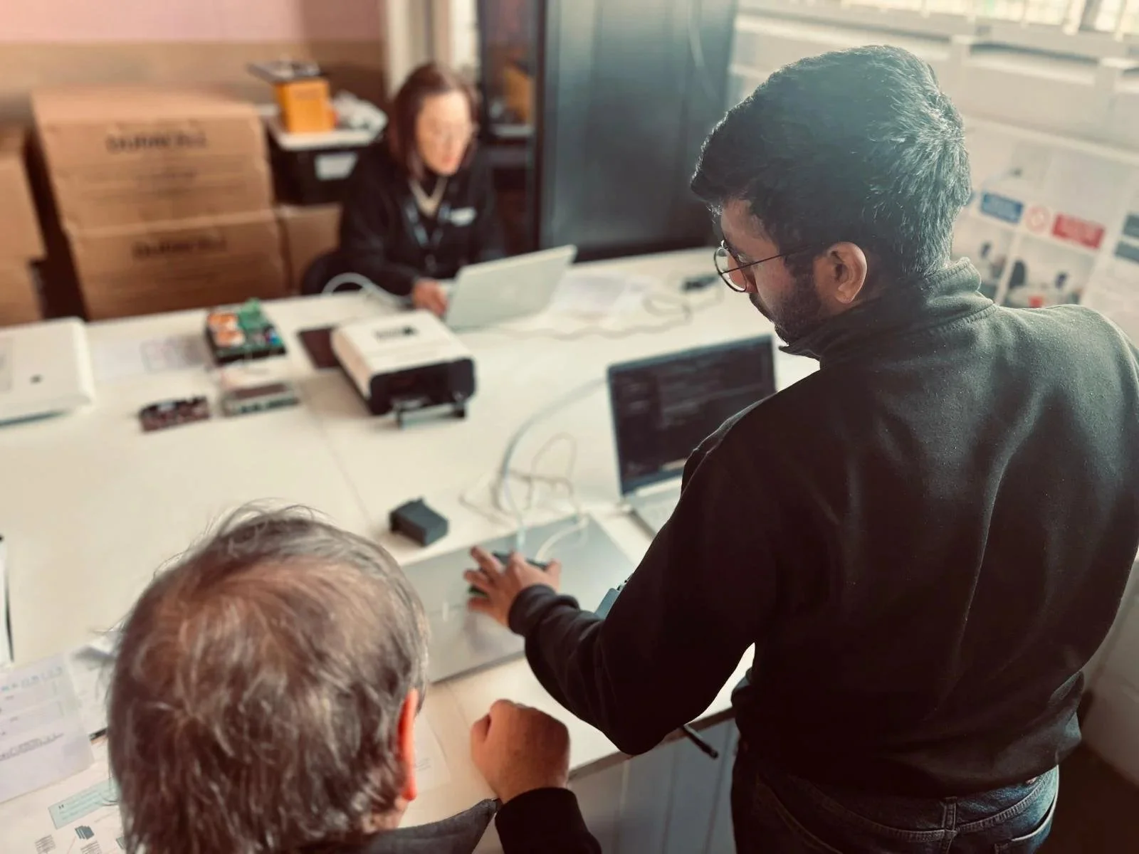 Three people gathered around a table in a product development room; two are sitting and one is standing, working on a laptop and reviewing specifications.