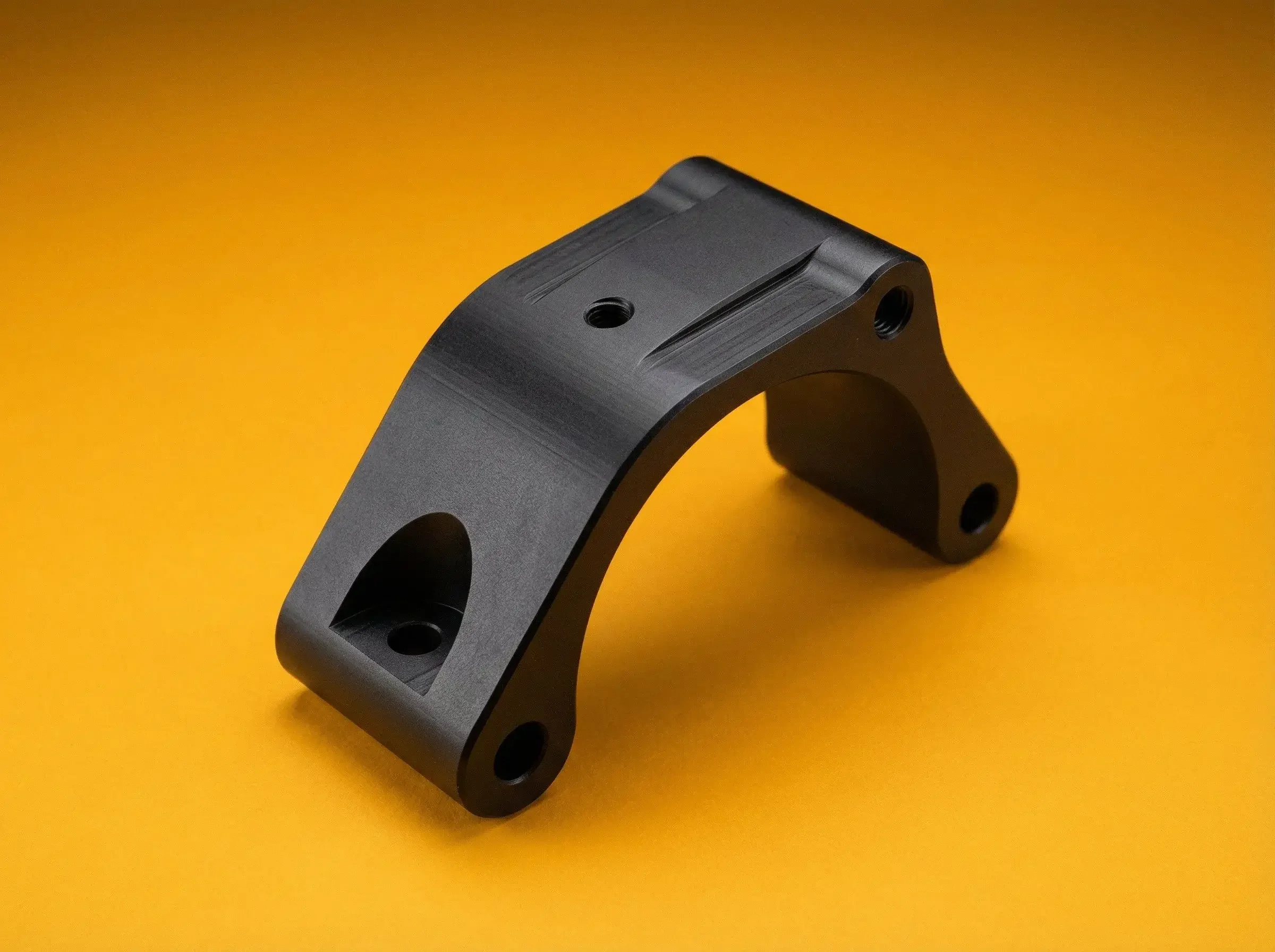 A black metal mechanical part with multiple holes, curved shape, and machined surface on a yellow background.