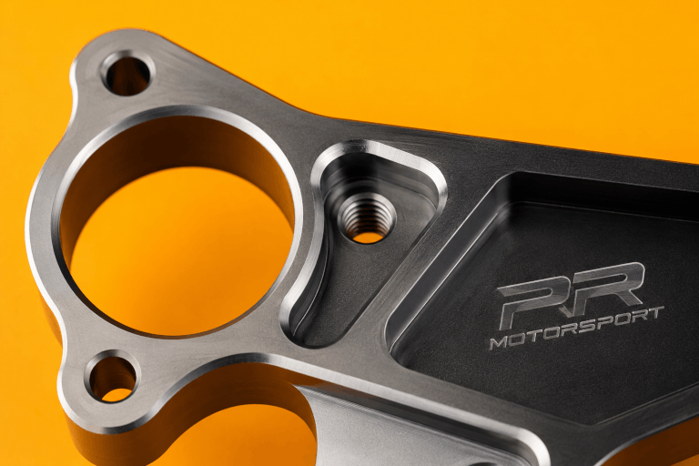Close-up of a black and silver car engine part with PR Motorsport logo on a yellow background.