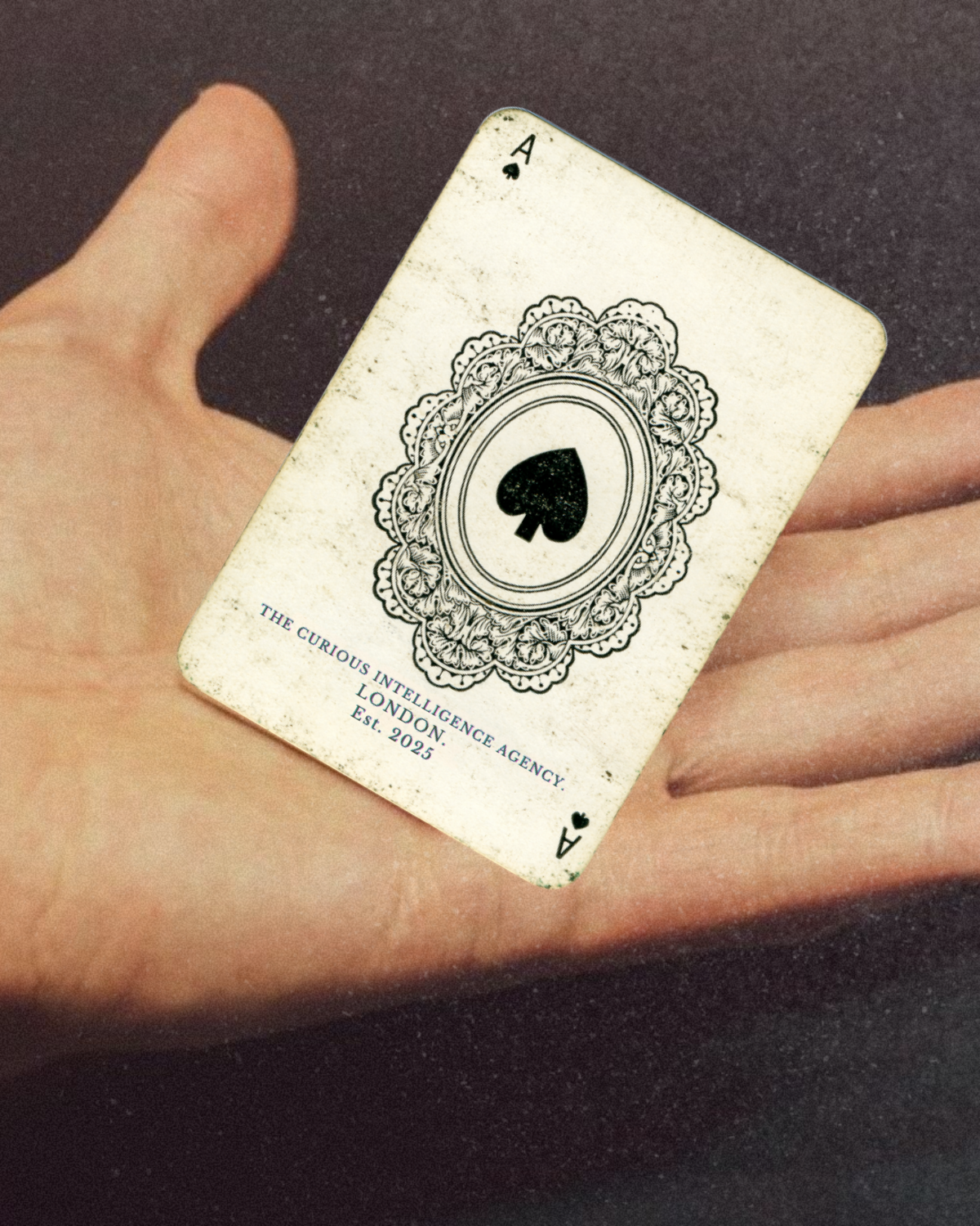 A hand holding a playing card, the Ace of Spades, with a ornate black and white design and a message about a fictional agency called The Curious Intelligence Agency, established in 2025.