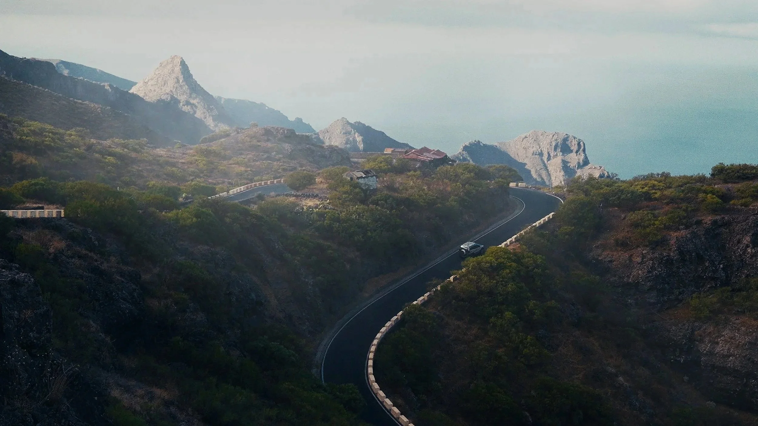 A winding mountain road with a black surface and white guardrails, surrounded by green and rocky terrain, with mountains in the background under a cloudy sky.