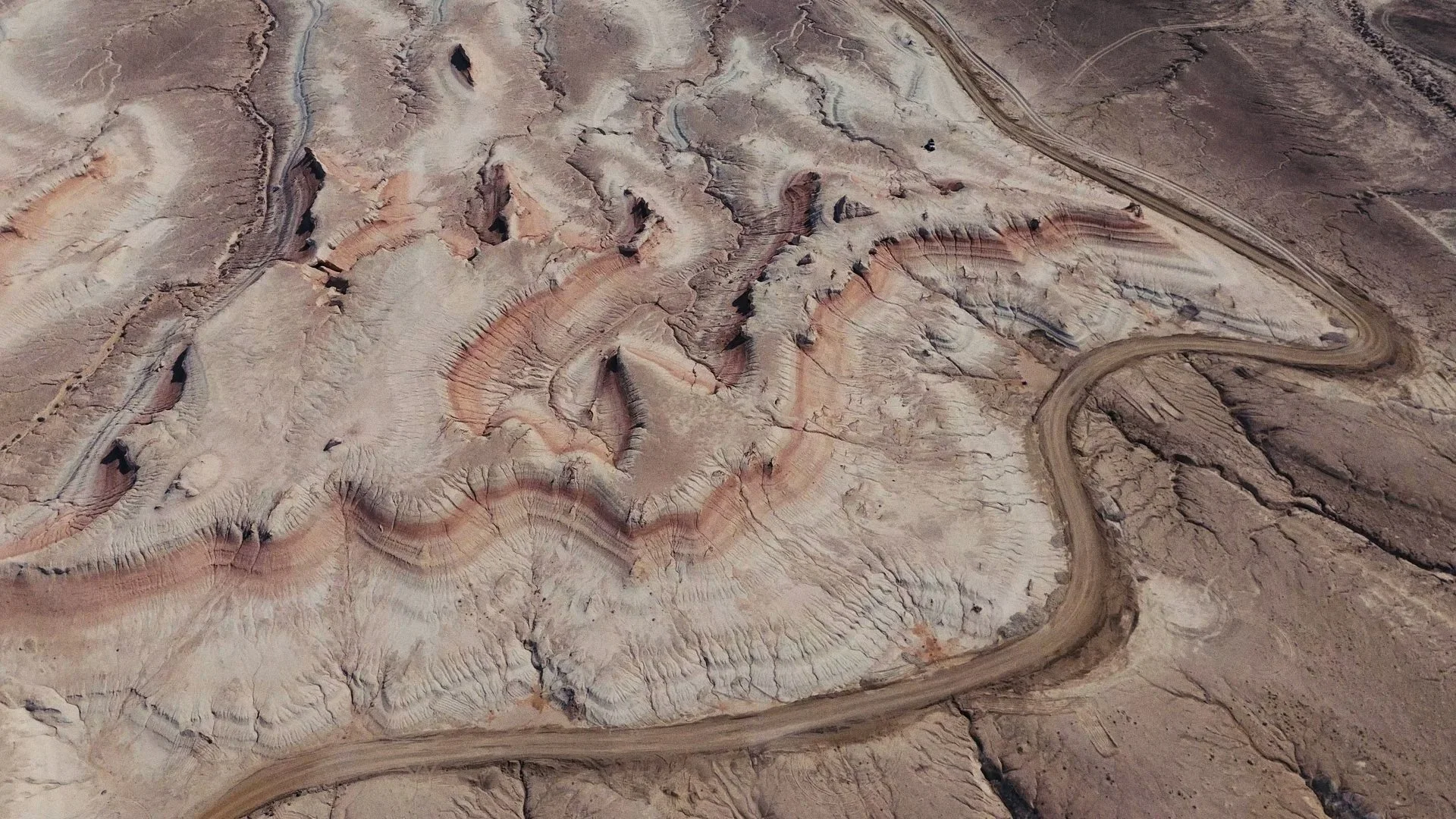 Aerial view of a desert landscape with rugged terrain, winding dirt road, and layered rock formations in shades of beige, pink, and brown.