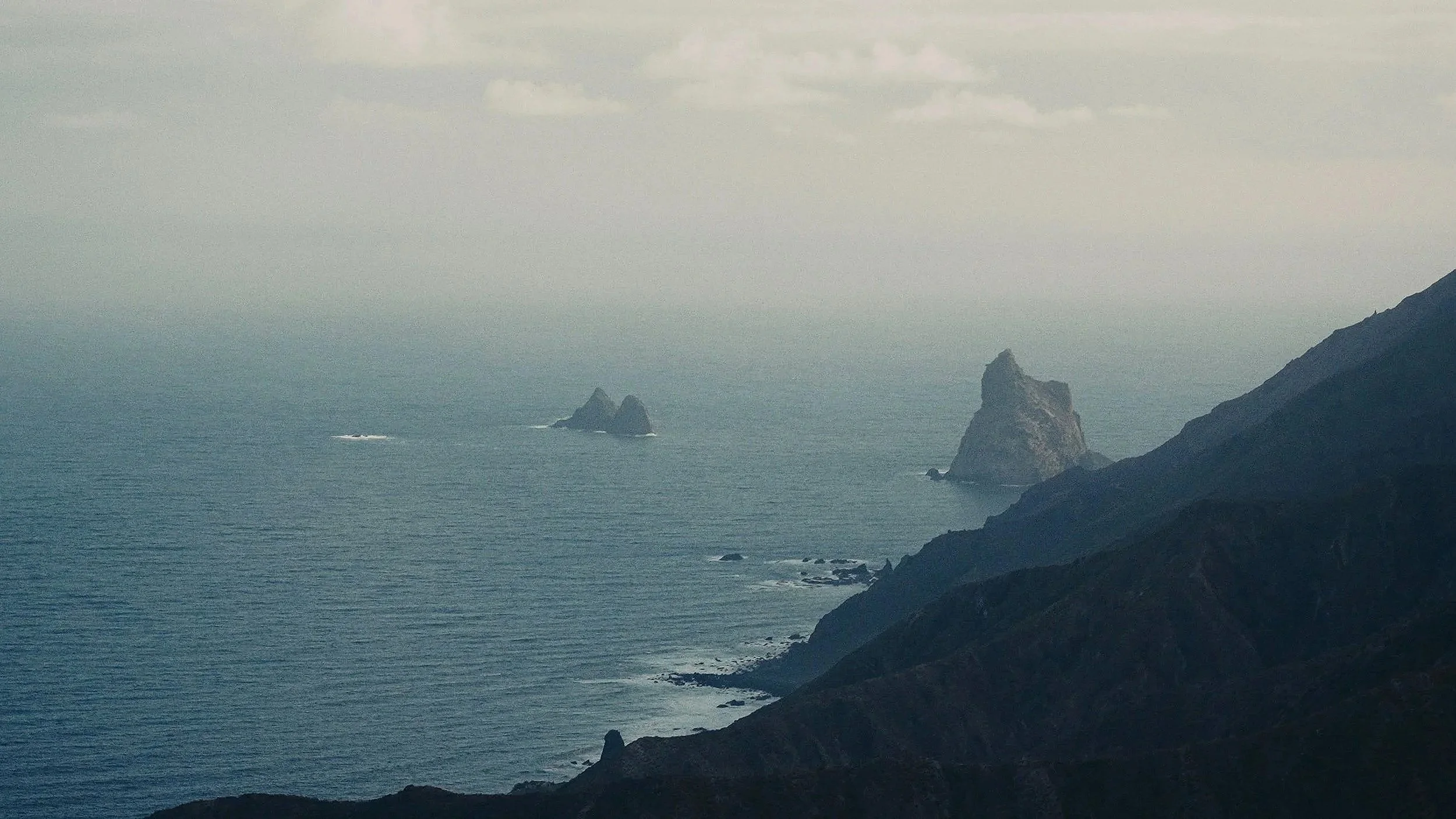 Coastal landscape with steep dark cliffs and rocky islands in the ocean under a cloudy sky.