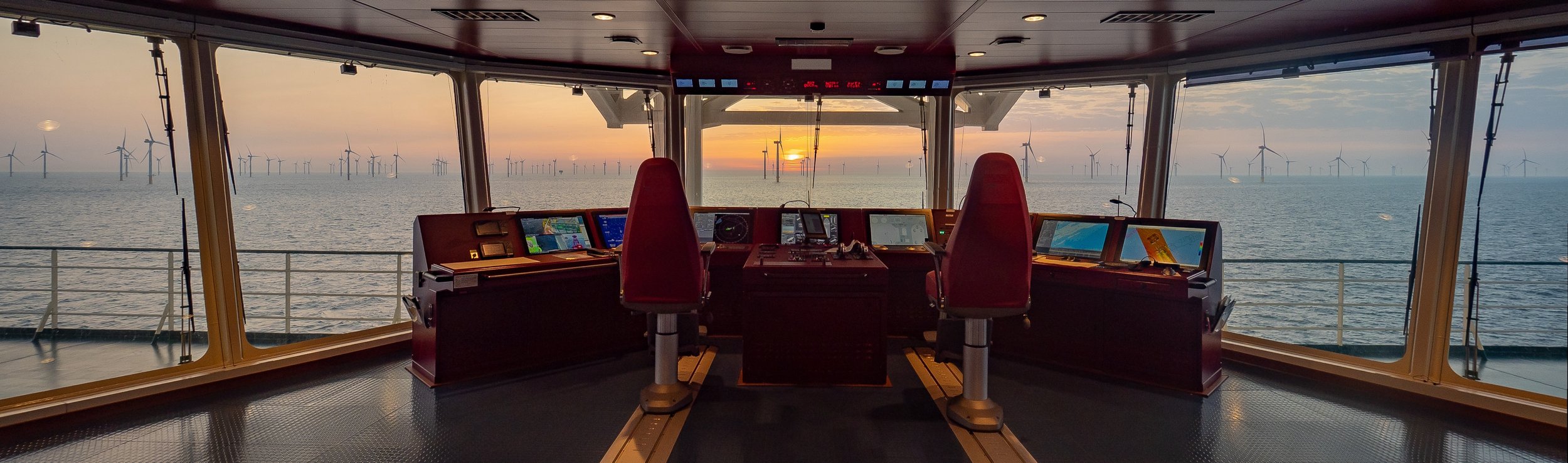 View from the ship's bridge showing navigation and control consoles with a sunset over an ocean and wind turbines in the background.