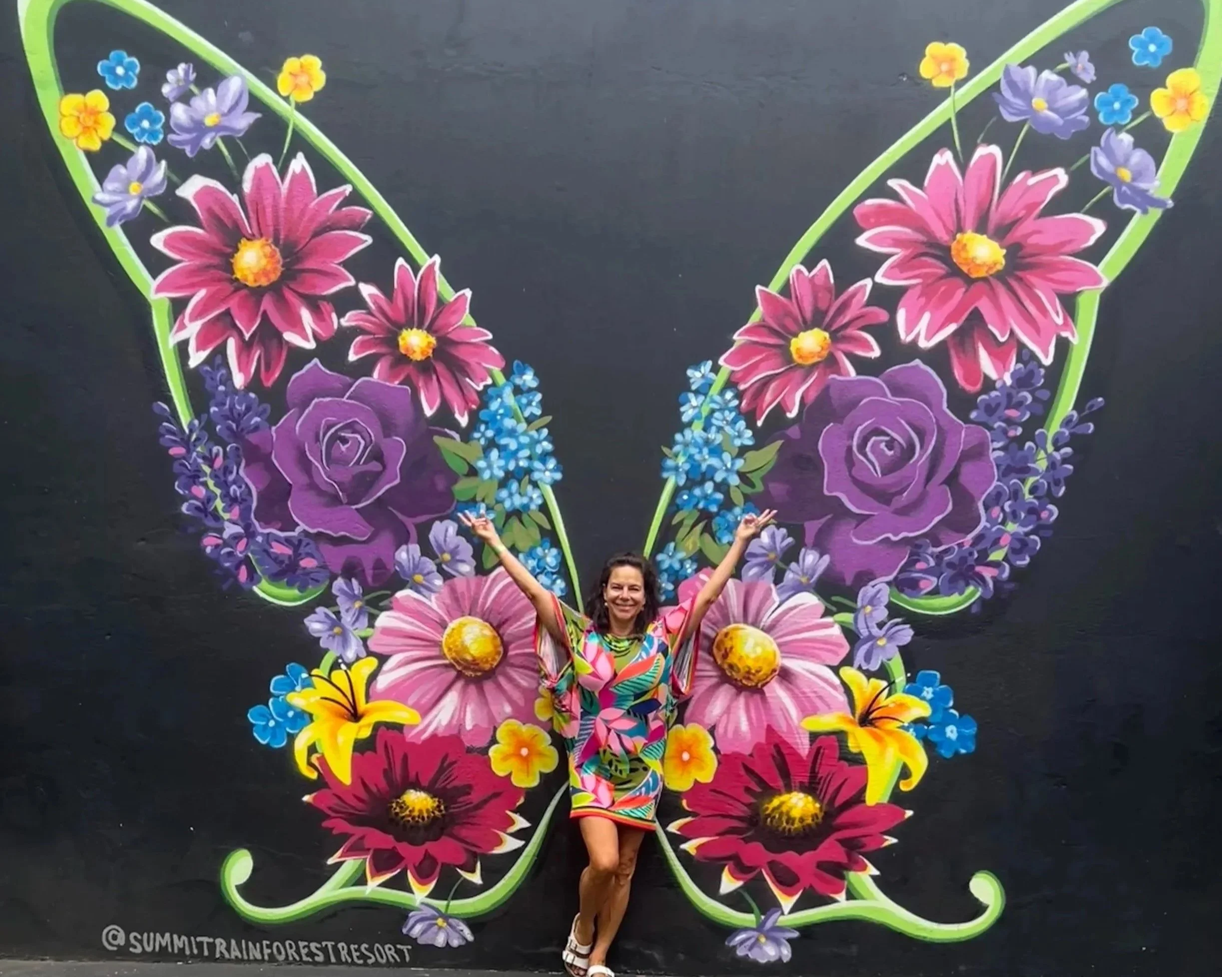 Woman standing in front of a large butterfly-shaped floral mural on a black wall, with arms raised in a joyful pose.