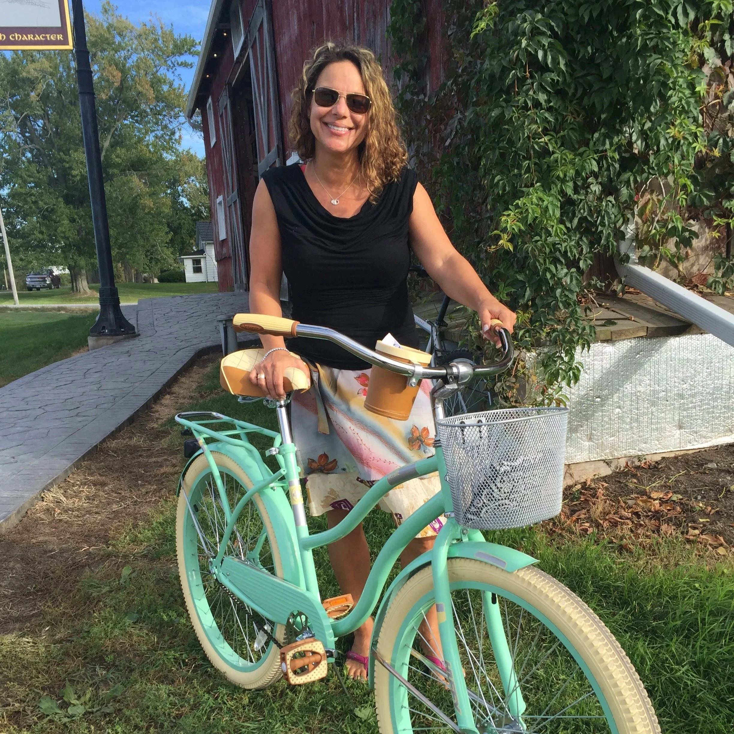 A woman with sunglasses and curly hair standing outdoors holding the handlebars of a mint green bicycle with a basket, in front of a red building with vines and trees in the background.