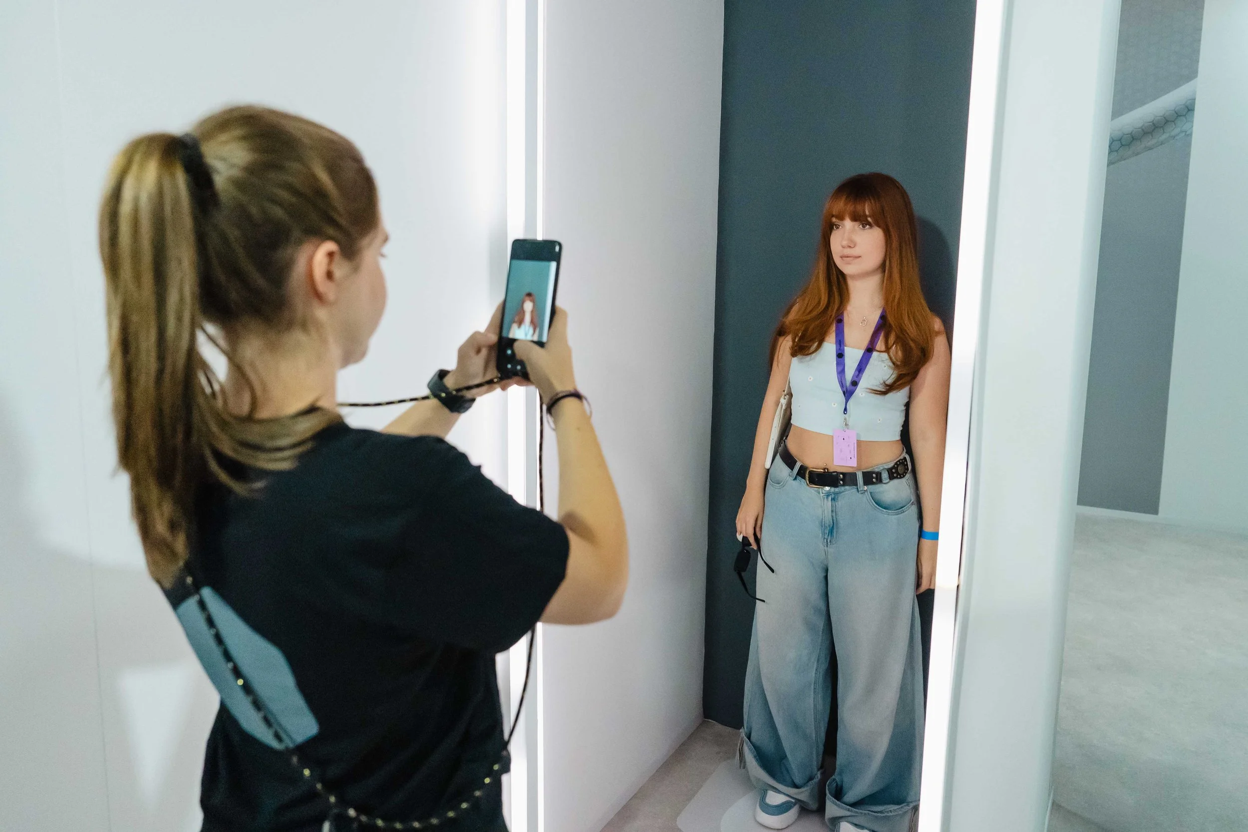 A woman with long red hair and a light blue cropped top stands in front of a mirror, posing for a photo taken by another woman who has long hair tied back. The person taking the photo is holding a cellphone and wearing a black shirt. The setting appears to be a modern, well-lit room with minimalist decor.