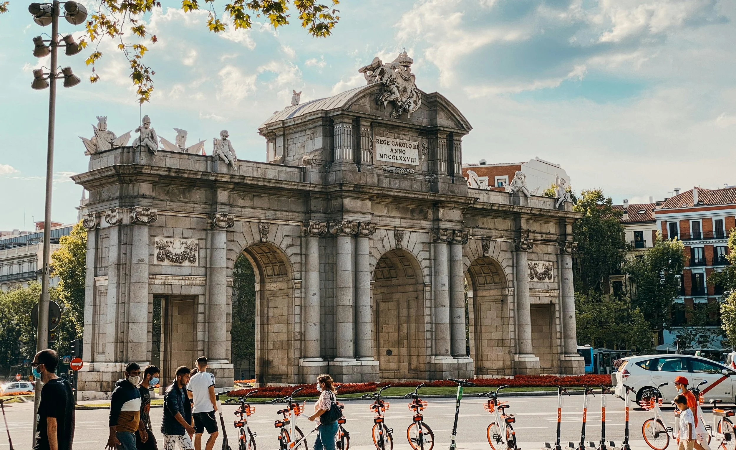 People walking past a historic arch monument with statues and a Latin inscription, in an urban area with bicycles, cars, and buildings around.