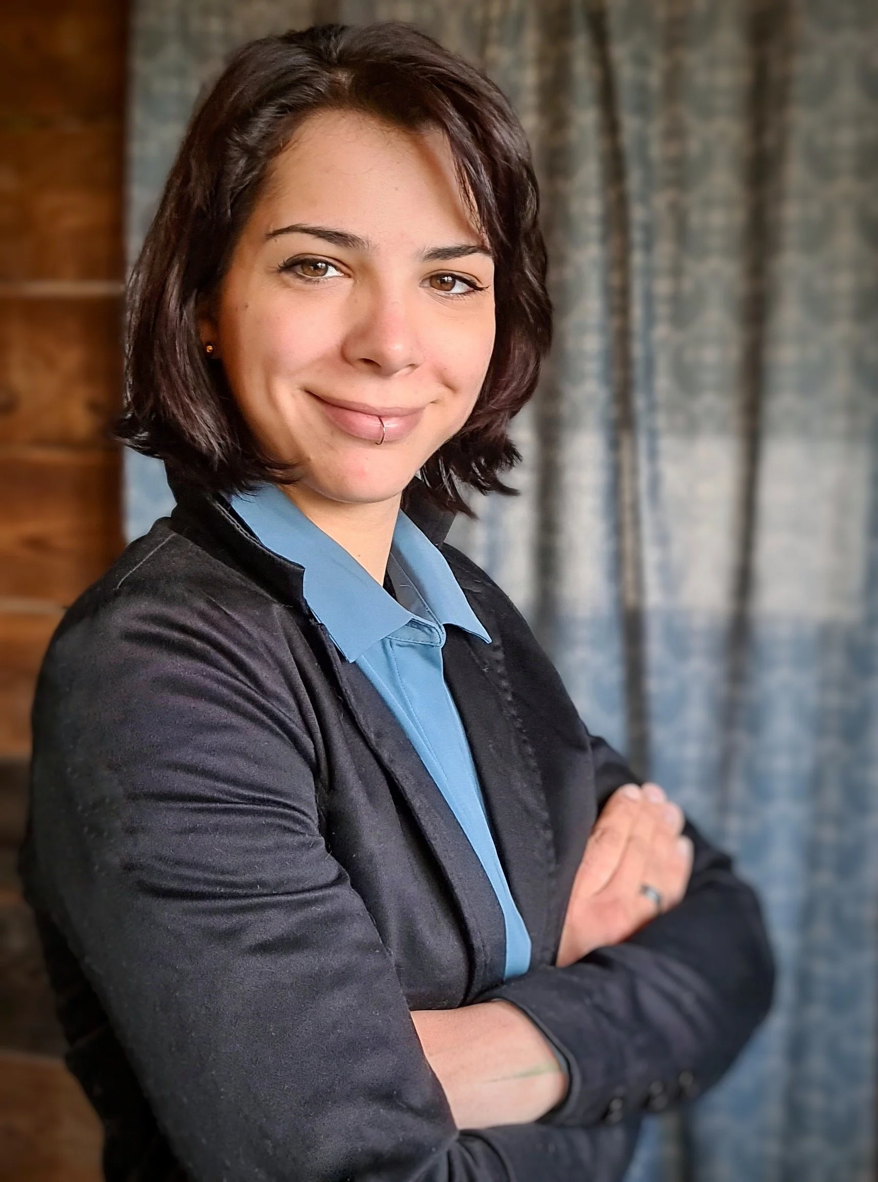 Ein junges Mädchen mit dunklen, schulterlangen Haaren, trägt eine schwarze Jacke und einen blauen Hemdkragen, steht vor einem Hintergrund aus Holz und Stoff, lächelt und hat Piercings an der Lippe.