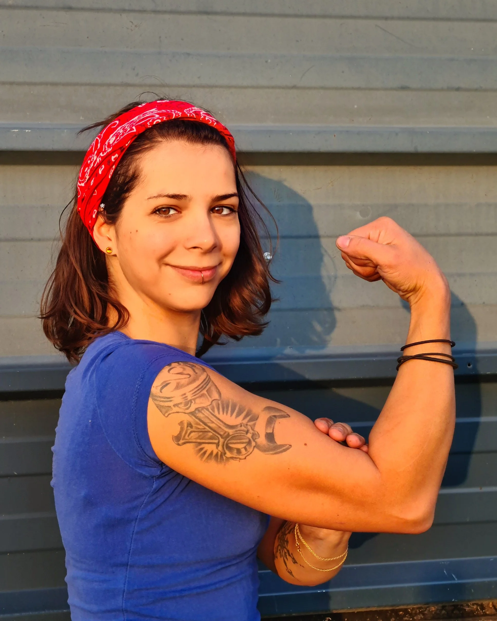 Frau mit Bizeps, roter Halstuch, blauer T-Shirt, Arm-Tattoo, wirft Muskeln und trägt Armbänder gegen den grauen Hintergrund