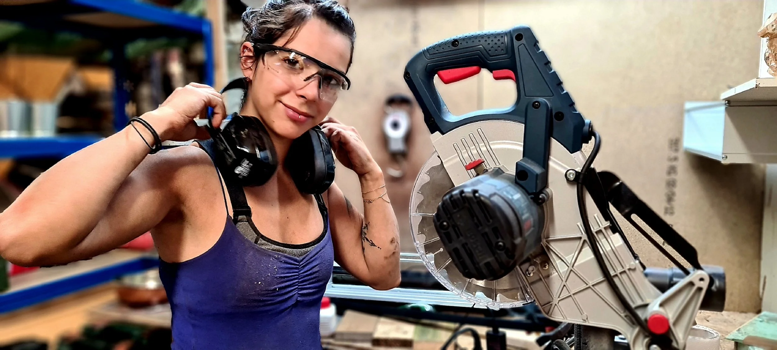Eine Frau in Schutzbrille und mit Gehörschutz in einer Werkstatt, die neben einer großen Handsäge steht.
