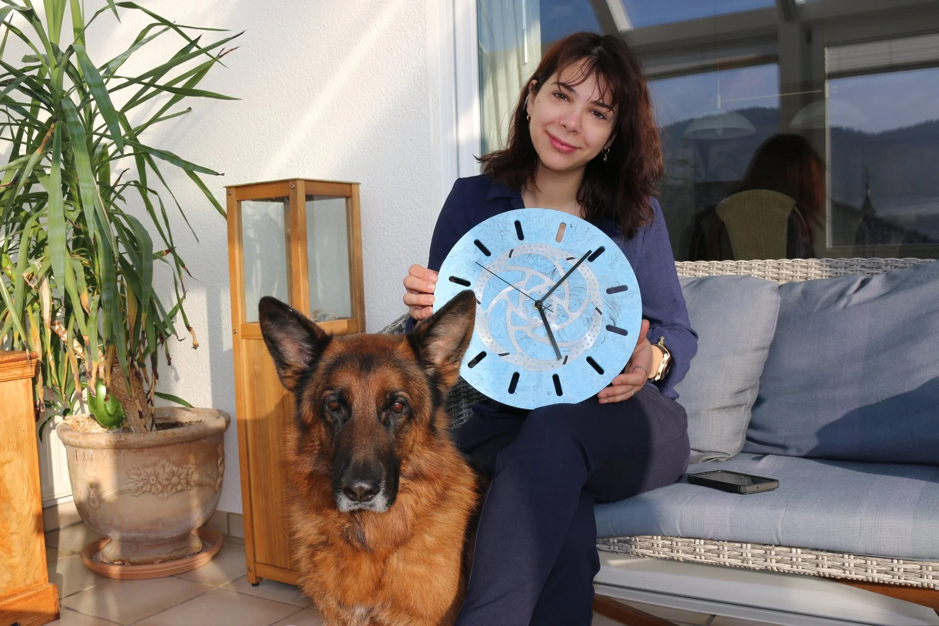 Eine Frau sitzt auf einer Terrasse mit einem Hund und hält eine blaue Mountenbike Fahrrad Wanduhr aus Jesmonite. Sie ist Handwerkerin und Luxemburgerin und bietet Handwerker Workshop für Frauen, Sie hat Ihr Label Creative little farm eröffnet