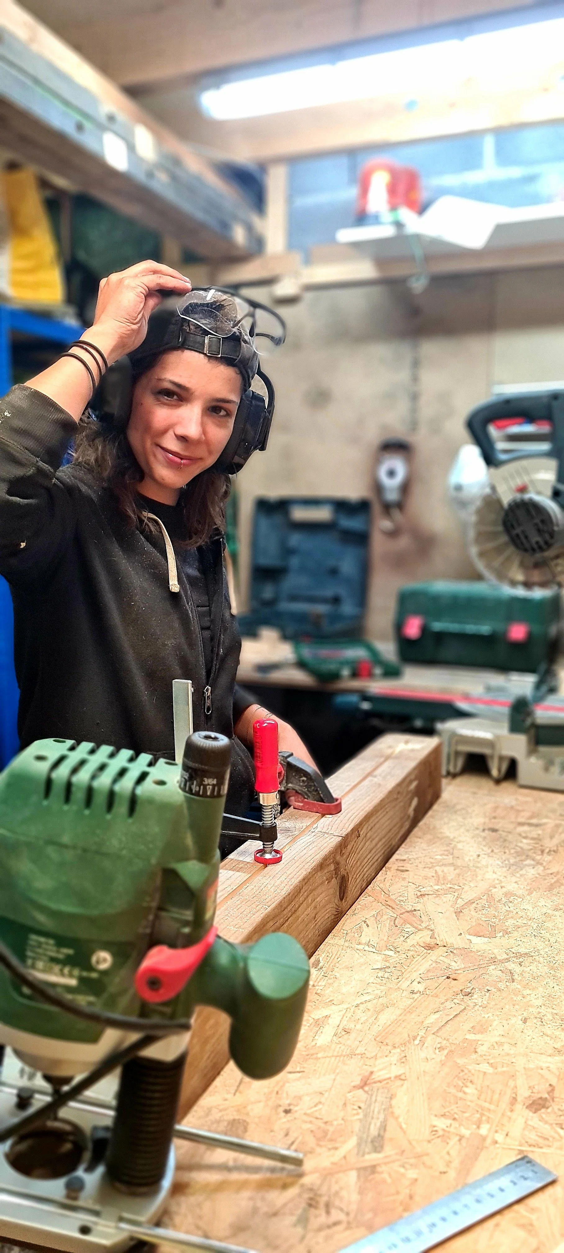 Eine junge Frau in einer Werkstatt trägt Gehörschutz und Schutzbrille, während sie an einem Holzstück arbeitet, umgeben von Werkzeugen und Baumaschinen. Sie ist Handwerkerin und Luxemburgerin und bietet Handwerker Workshop für Frauen