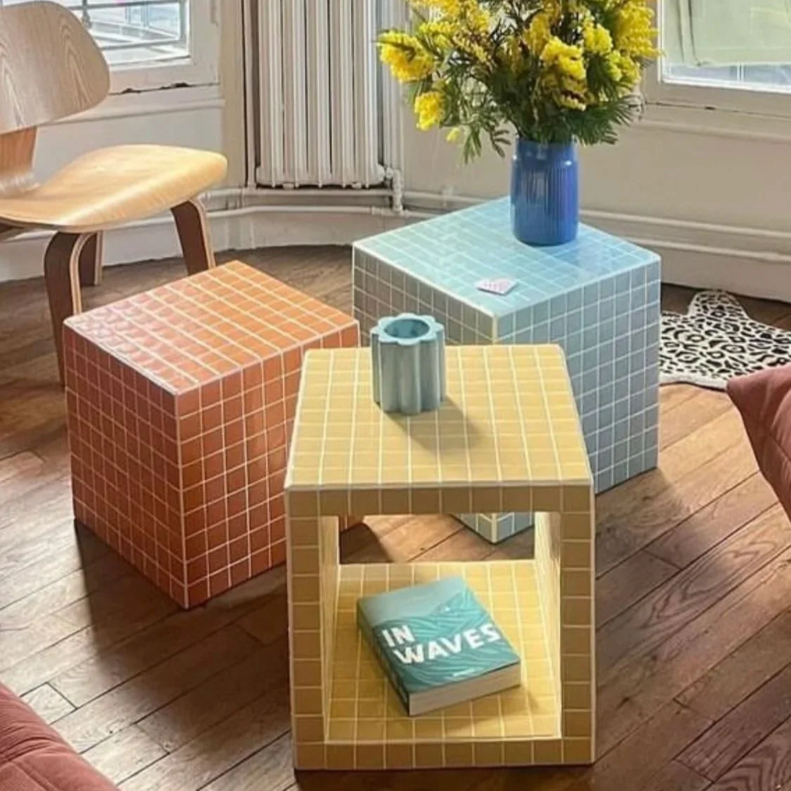 Drei quadratische und bunte Möbelstücke mit Fliesenmuster, in der Mitte eine Vase mit gelben Blumen, auf den Tischen liegen Gegenstände, in einem Raum mit Holzfußboden und Fenstern im Hintergrund. Handwerker Workshop für Frauen