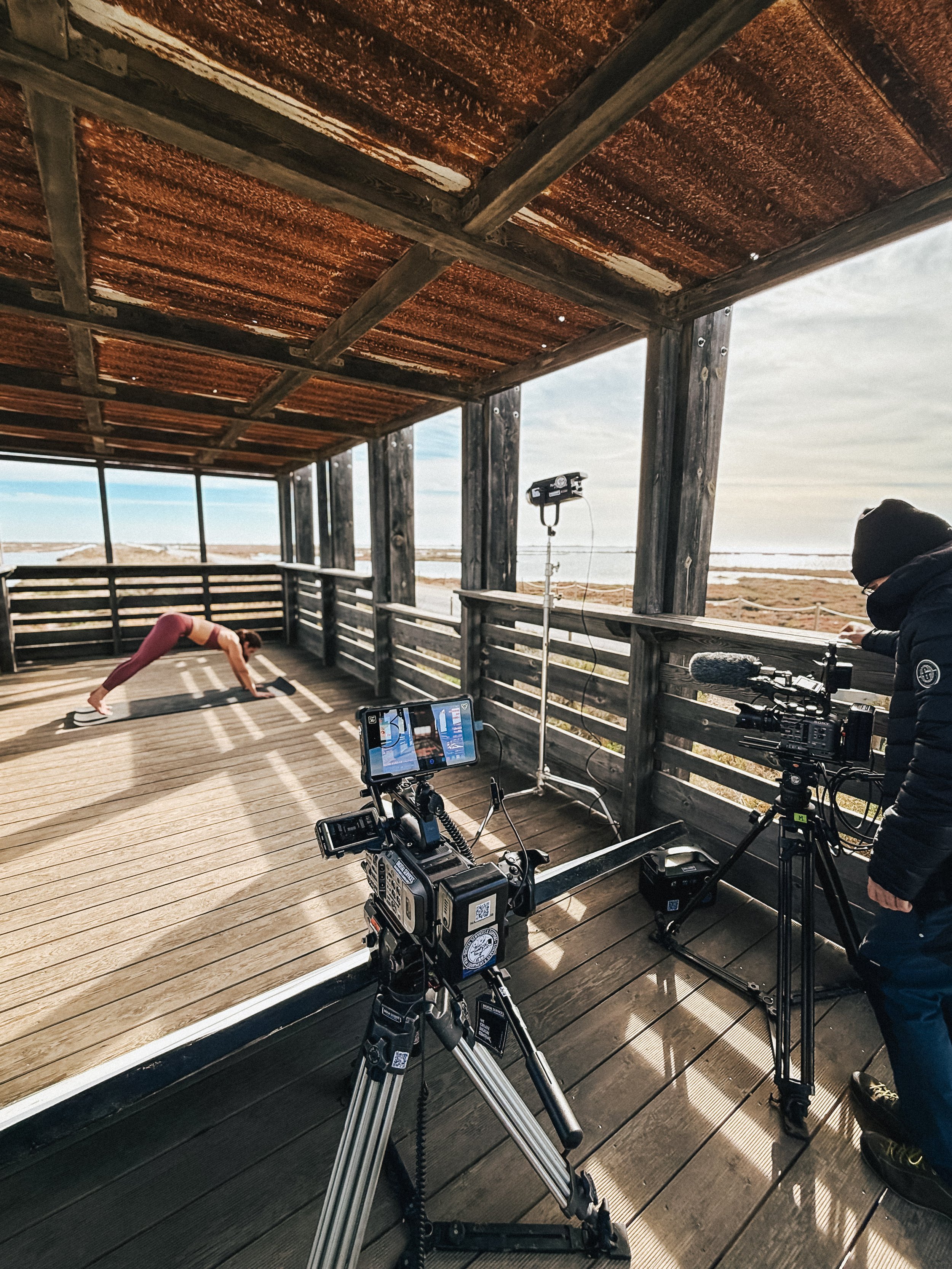 A woman is performing yoga on a wooden deck under a rustic roof while a camera crew films her from behind.