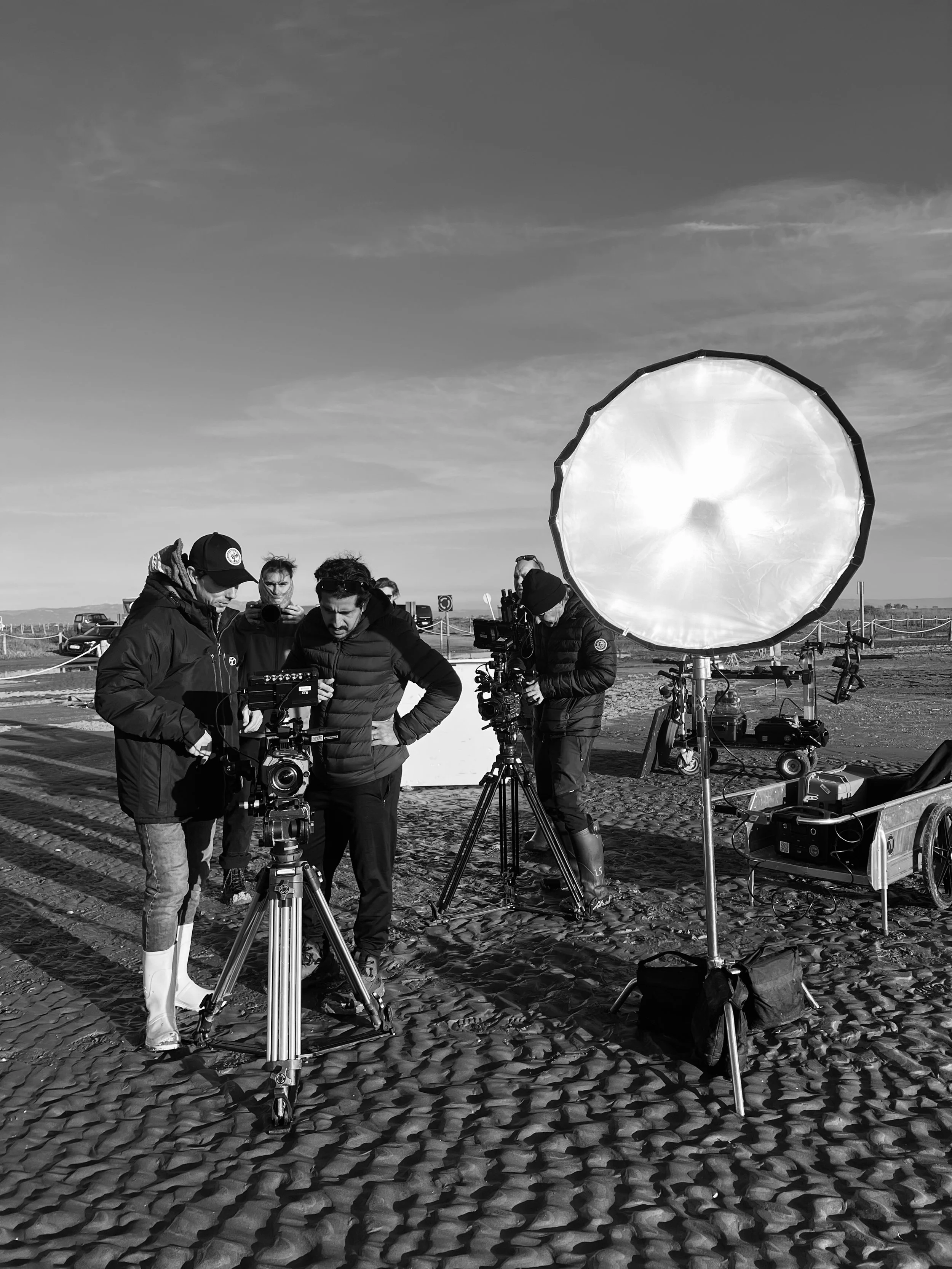 Film crew preparing equipment for outdoor filming on a sandy location, with large lighting equipment and cameras.