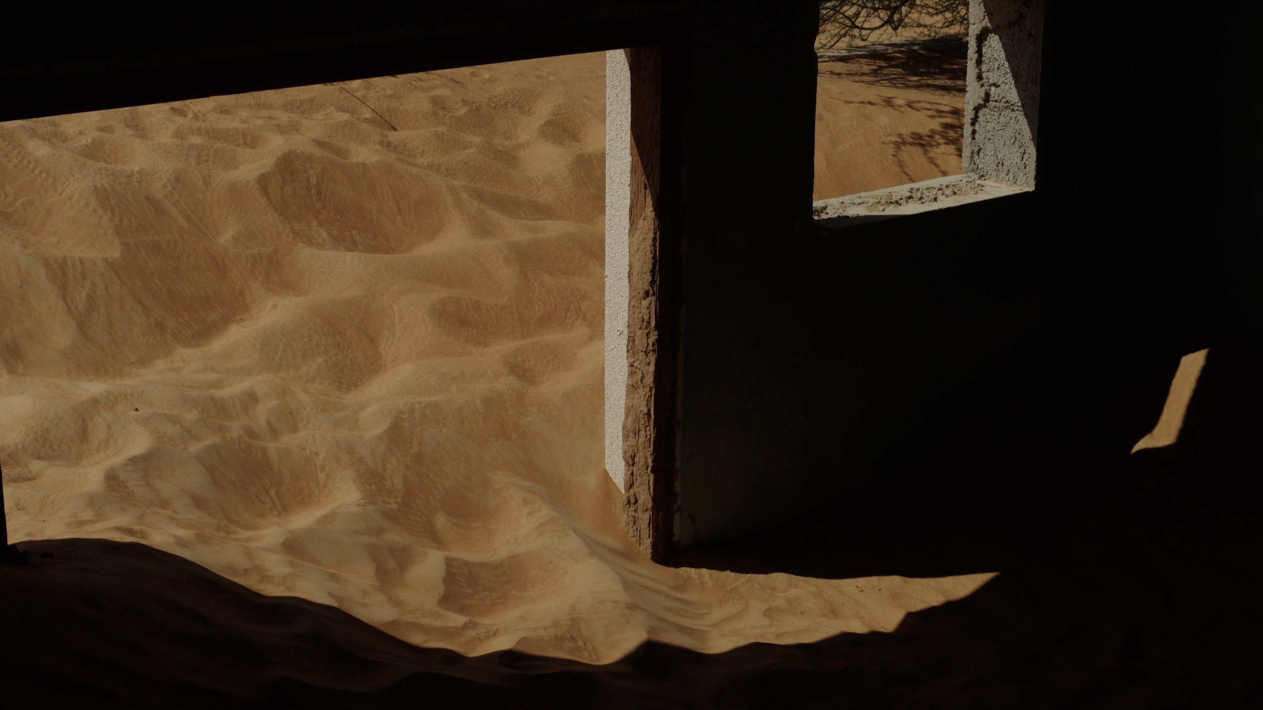 View through a small rectangular window in a desert structure showing sandy dunes outside with shadows cast on the interior wall.