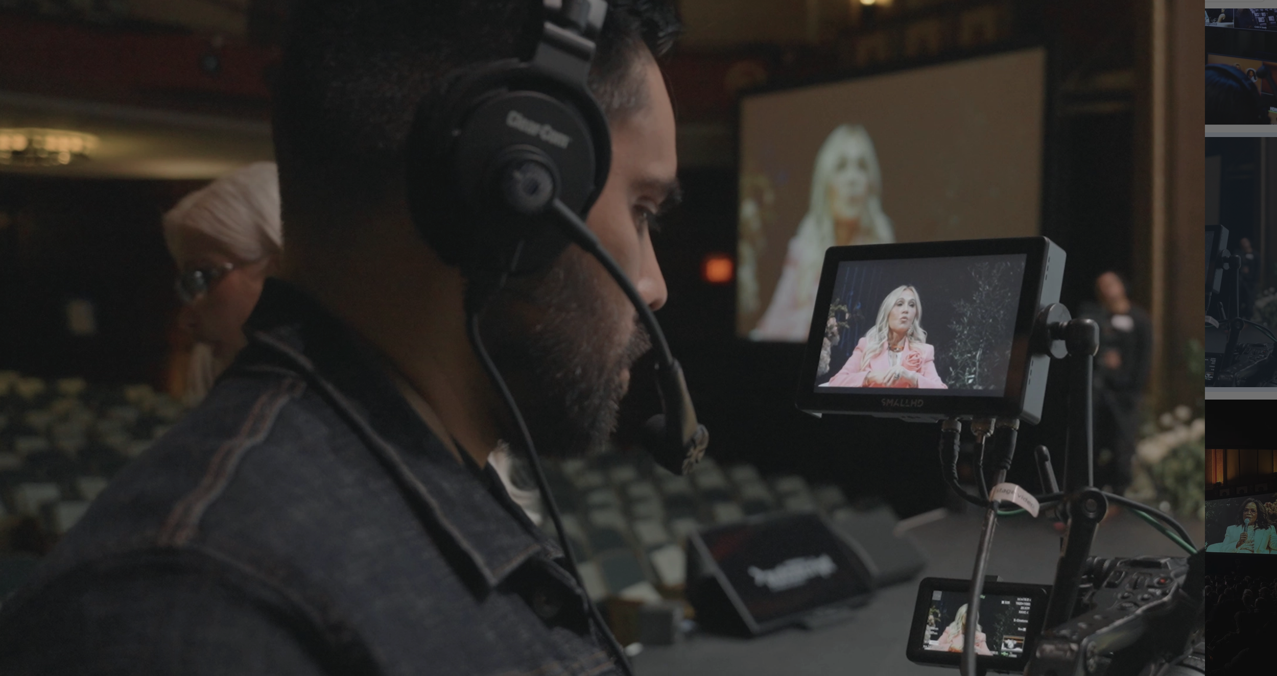 A man operating a camera with a mounted monitor during a live event, with a woman in pink on stage projected on a large screen behind him, and audience seats visible in the background.