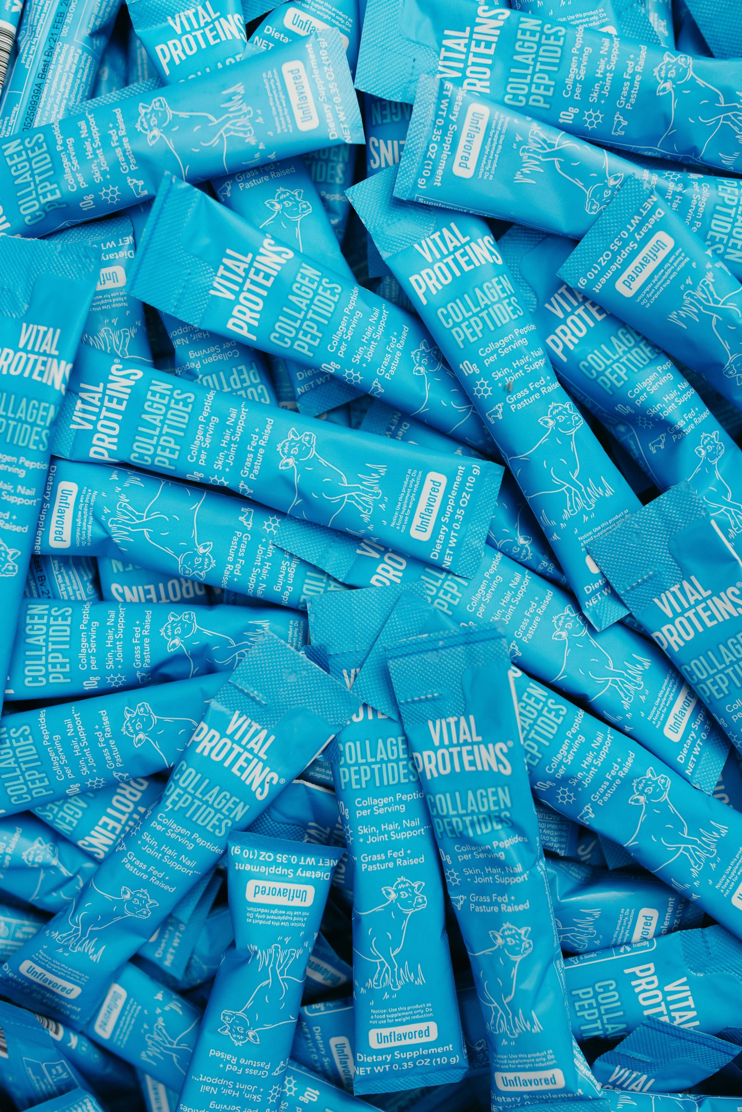 Pile of blue dietary supplement packages labeled 'Vital Proteins Collagen Peptides' with a cow illustration, indicating unflavored collagen supplement for skin, hair, nails, joint support, and grass-fed pasture-raised.