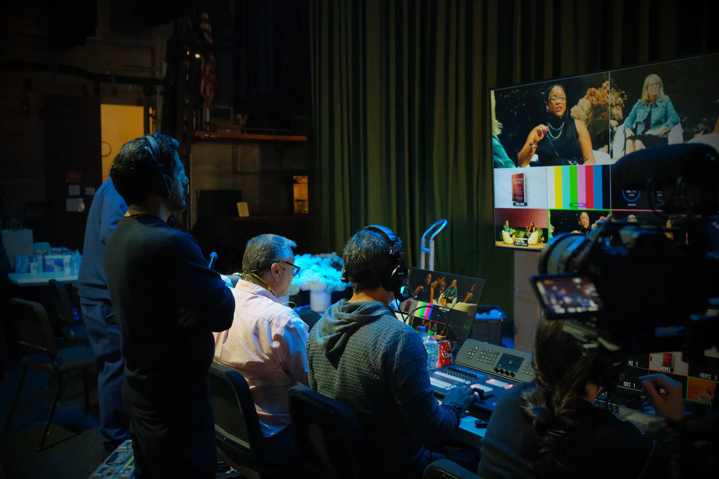 Television production crew working in a studio, with some people operating cameras and equipment while watching a large screen displaying multiple video feeds of a woman speaking during a panel discussion.