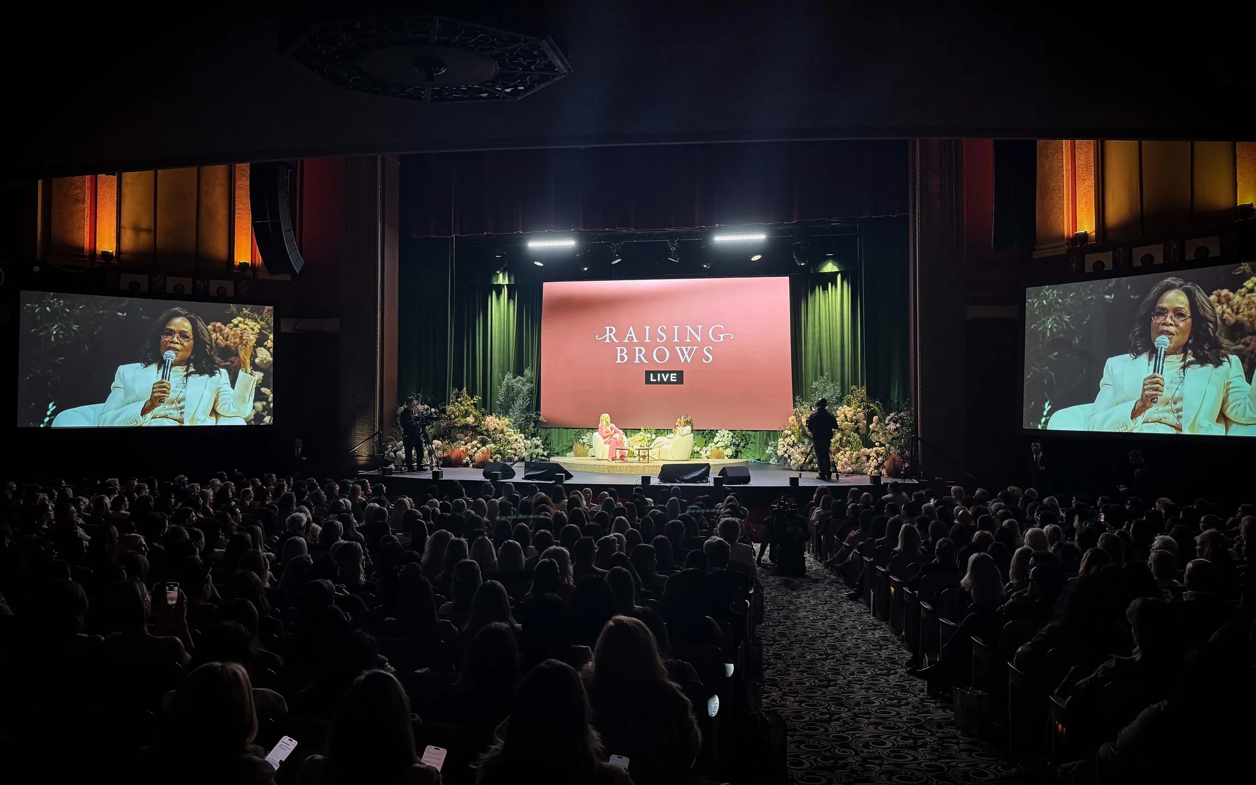 A large audience watching a live panel discussion titled "Raising Brows" on stage with two women seated in chairs, floral arrangements, and large screens displaying a woman speaking into a microphone.