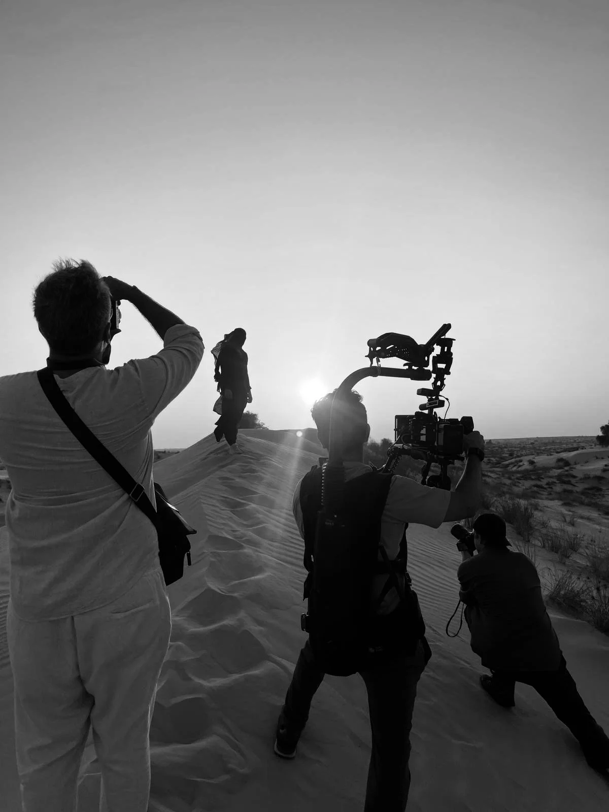 Filmmakers filming a scene in a desert at sunset, with one person standing on sand dunes and others operating camera equipment.